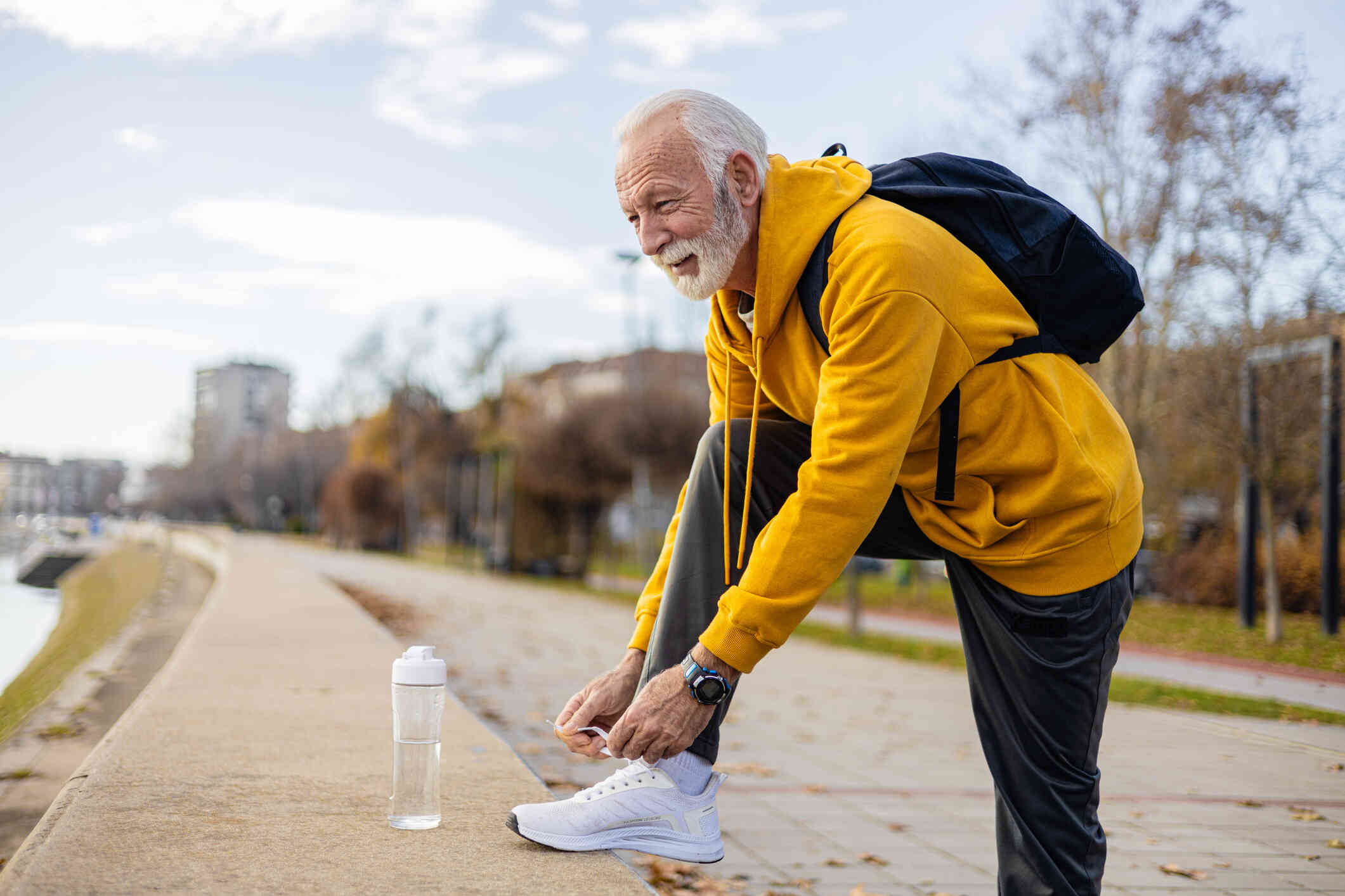 A mature man wearing a yellow hoodie and a backpack props his foot up on a small concrete wall to tie his shoe as he stands outside on a walking path.