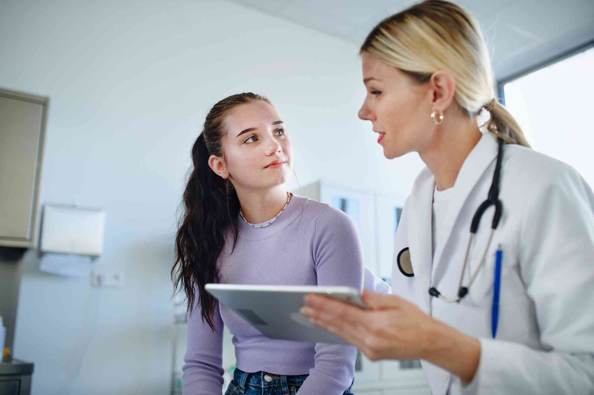 A teen girl in a purple shirt sits on an exam table while talking with a woman in a white coat holding a tablet