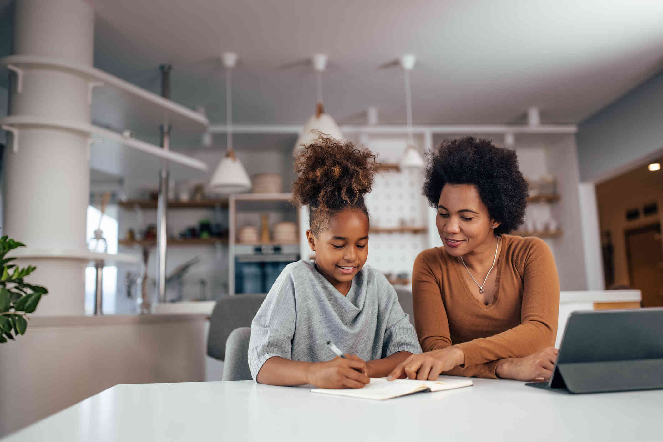 A mother sits at the kitchen table with her daughter and helps her with her homework by pointing to information in a book.