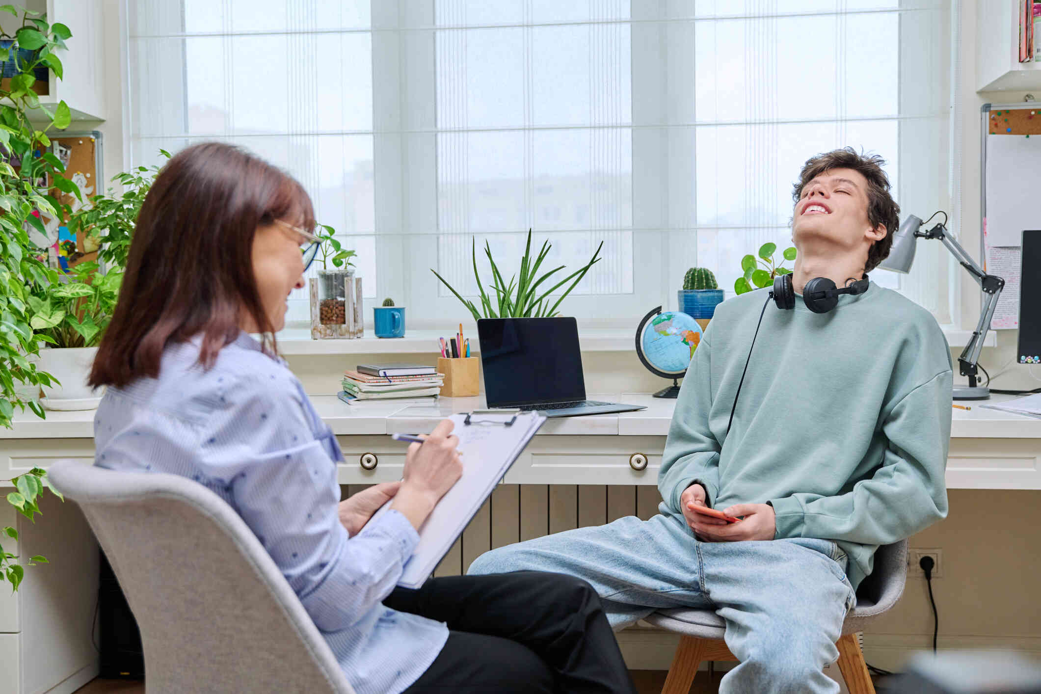 A young man with headphones around his neck leans his head back smiling while sitting in an office across from an older woman marking things down on a clipboard.