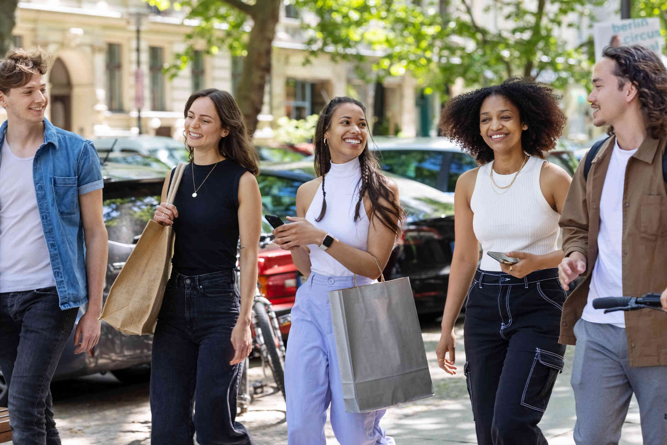 A group of adult friends walk along the side walk together on a sunny day while laughing and chatting.