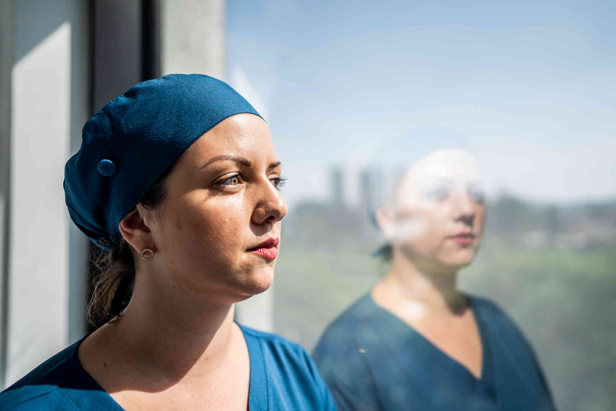 A female surgeon in blue scrubs stands near a window and gazes out while deep in thought.
