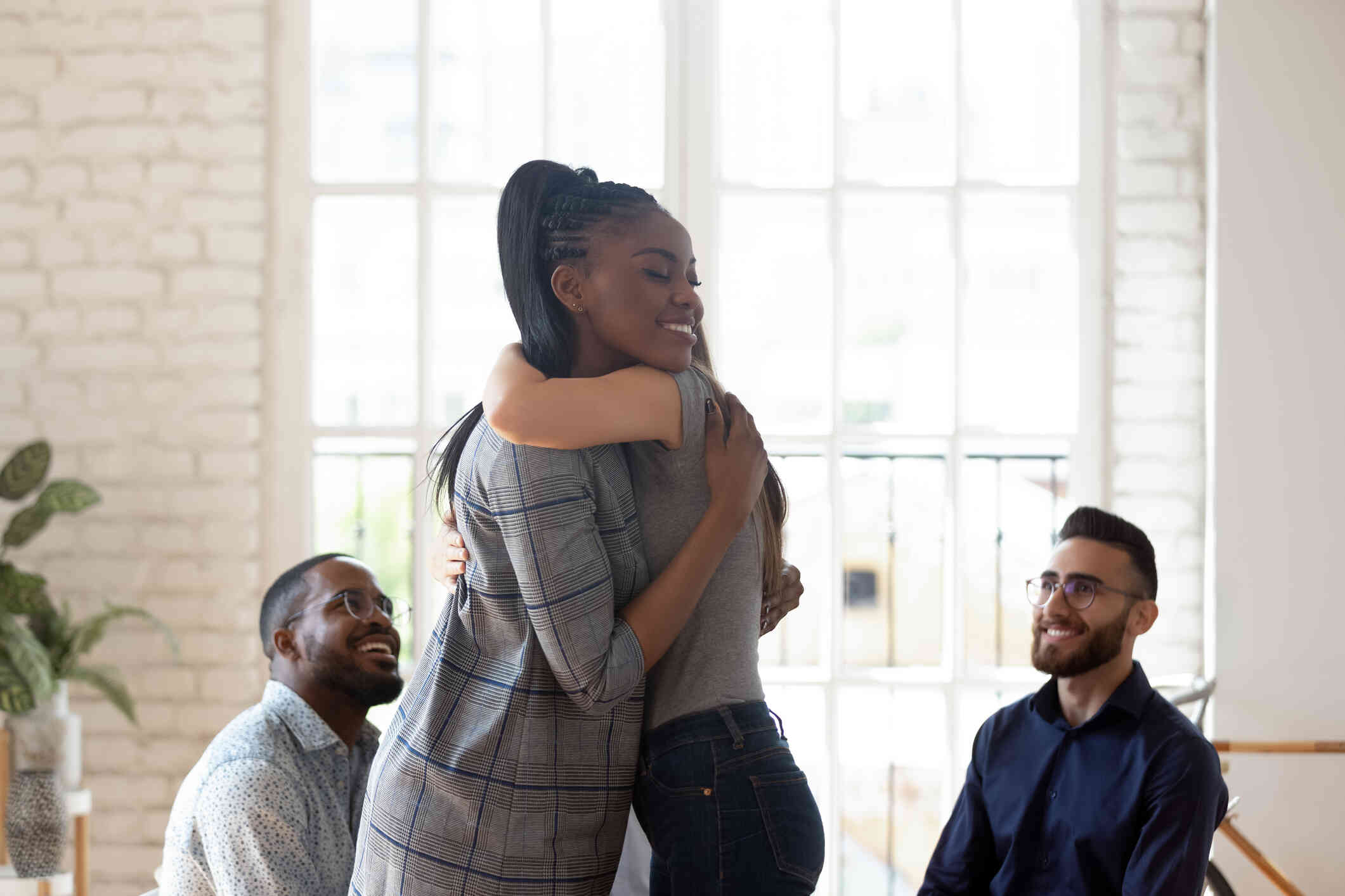 A woman in a blazer smiles as she hugs another woman in a room with large windows. Two men sit in chairs behind them and smile.