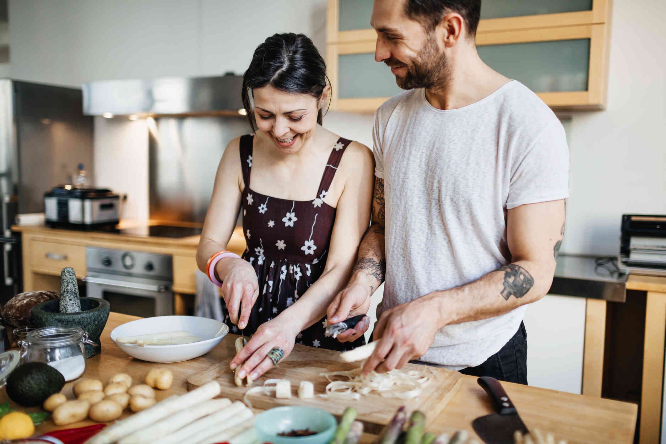 A male and female couple stand side by side in the kitchen and chop vegitables together while smiling.