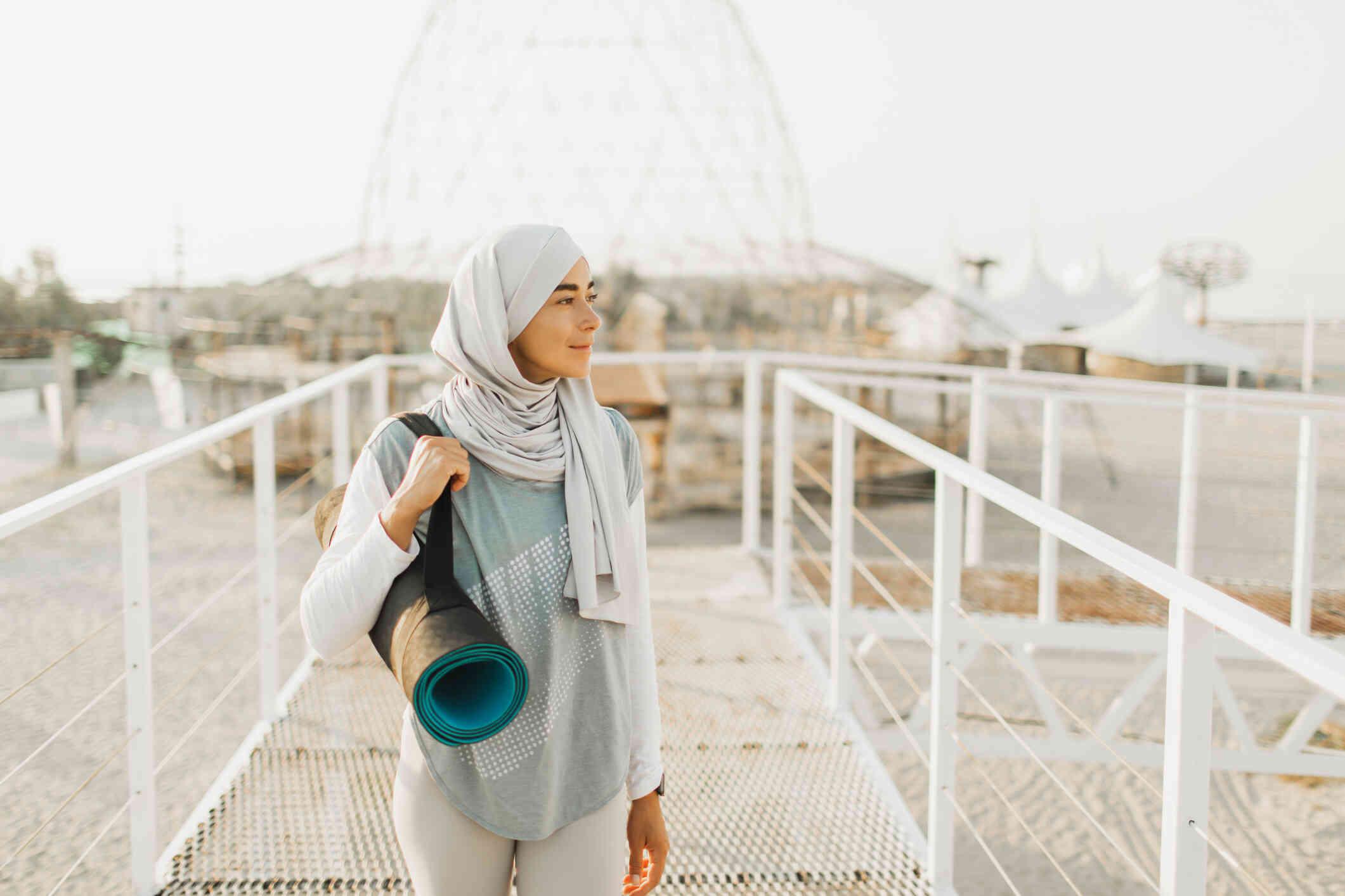 A woman wearing a hijab walks along a bridge while holding a yoga mat and glazing off.