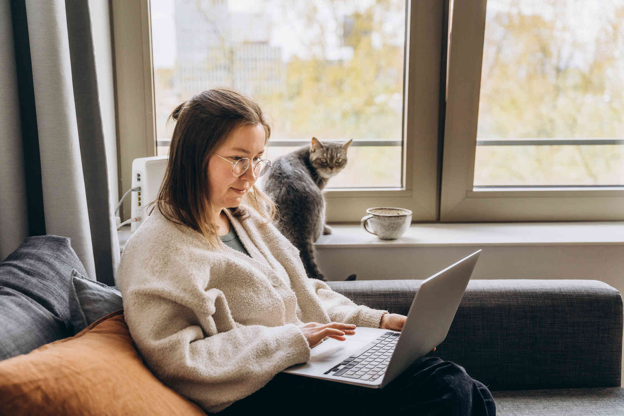 A woman in a tan sweater sits in a chair next to a window with a cup of tea and a cat while typing on the laptop in her lap.