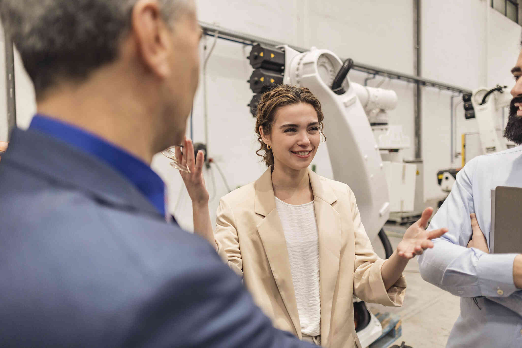 A woman in a tan blazer smiles as she stands in front of machinery in a warehouse and speaks to two men beside her.