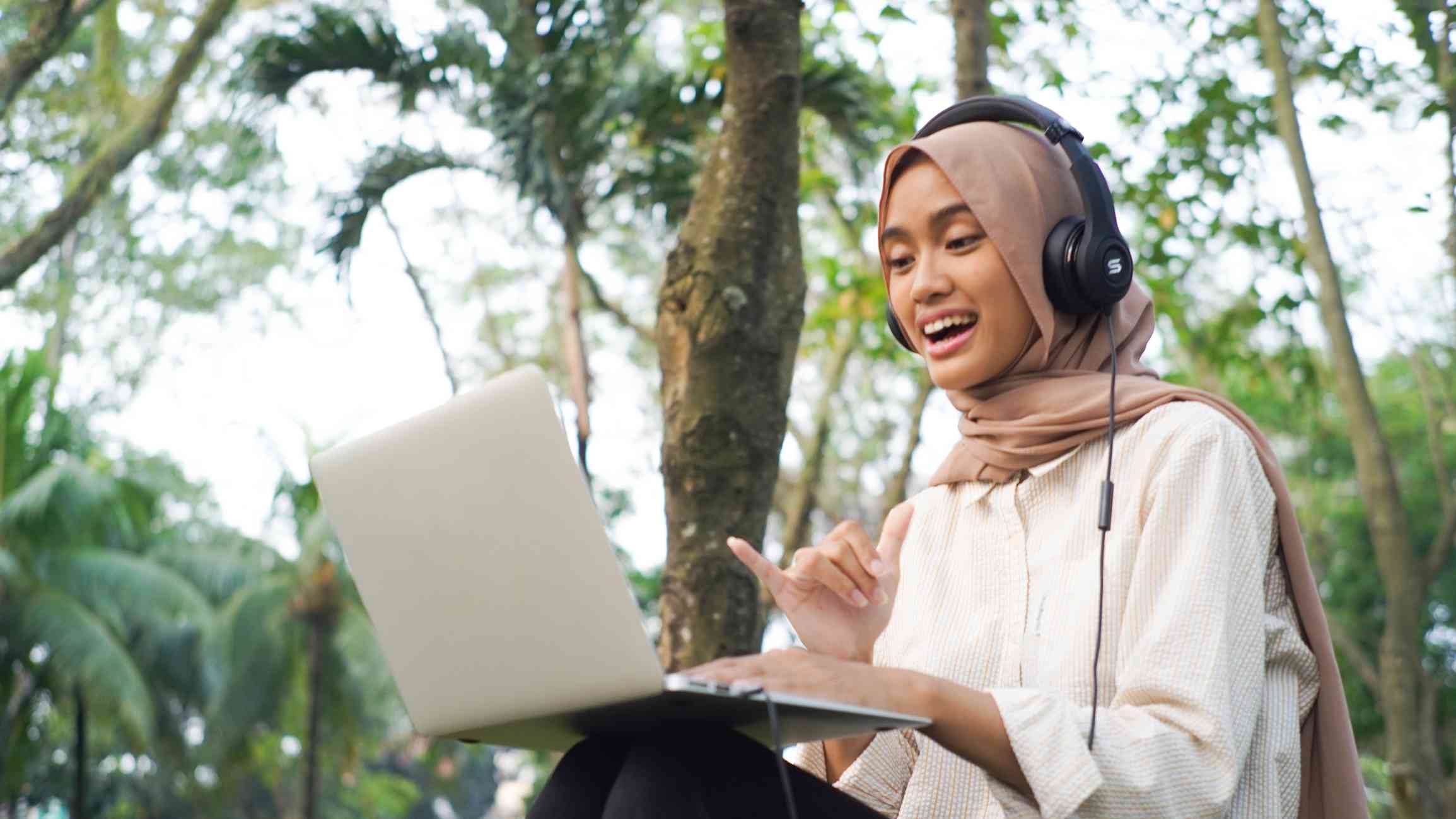 A woman in a pink hijab sits outside at a table with headphones on as she smiles and signs to the open laptop infront of her.