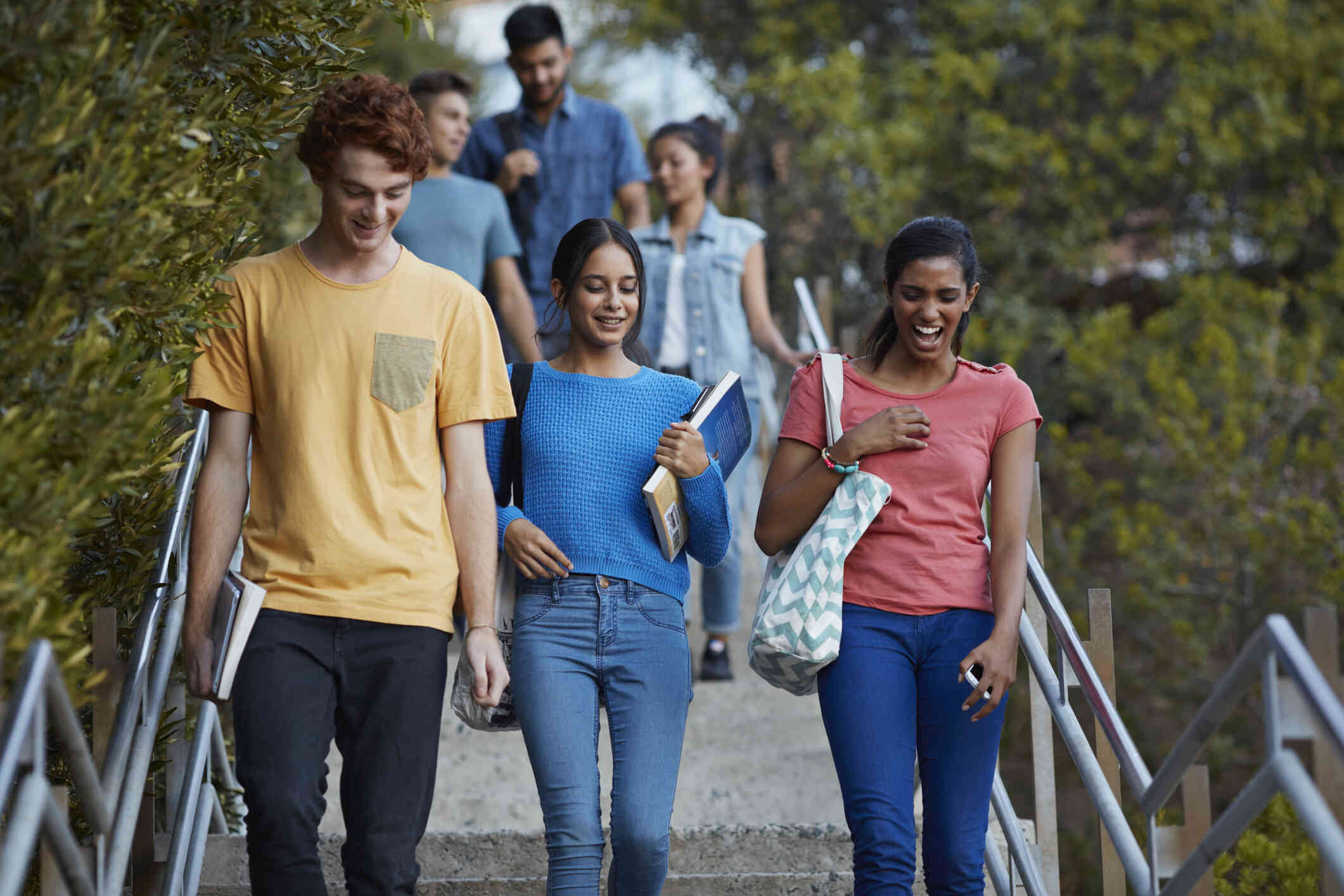 Three college students walk down a staircase on campus. One man in a yellow shirt looks down, and two women carrying books and a tote bag laugh beside him.