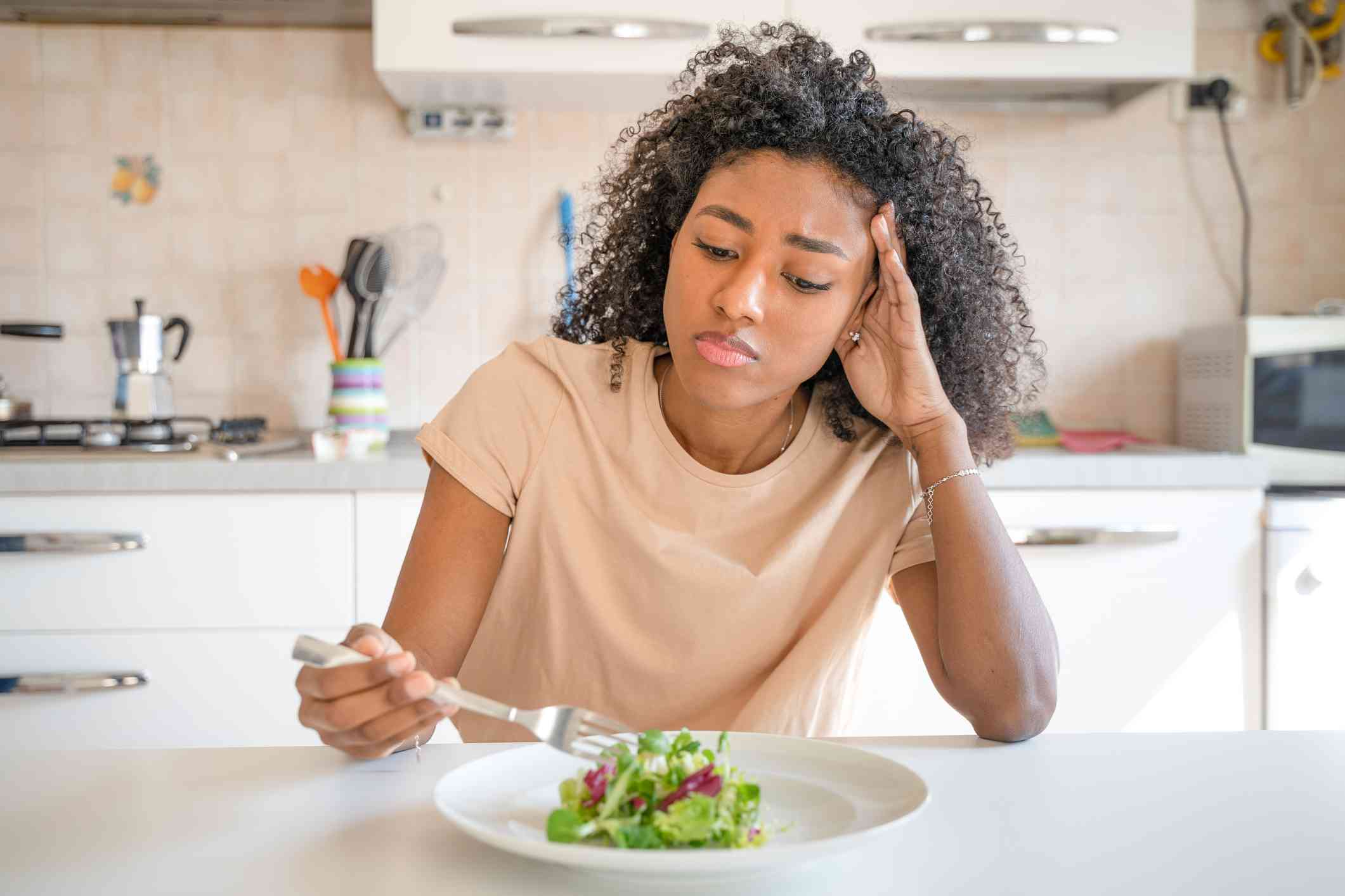A woman in a tan shirt sits hunched over a salad at the kitchen table with a sad epression as she picks at the food.