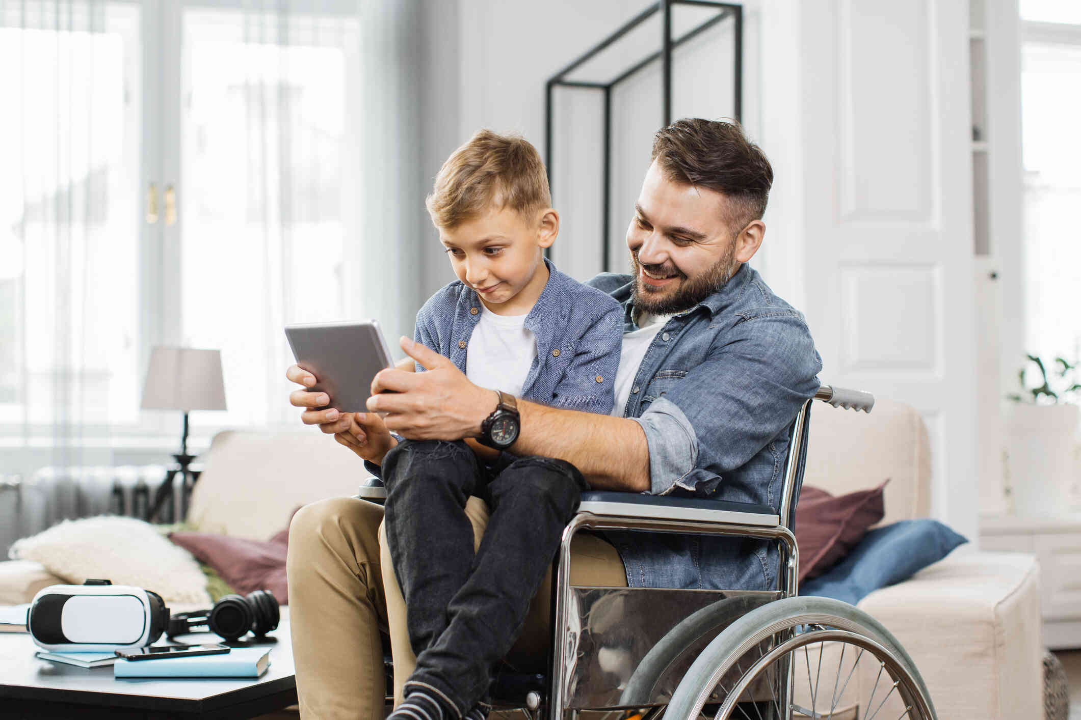 A boy sits on his fathers lap as he sits in a wheelchair in the living room and they both look at the tablet in the fathers hands.