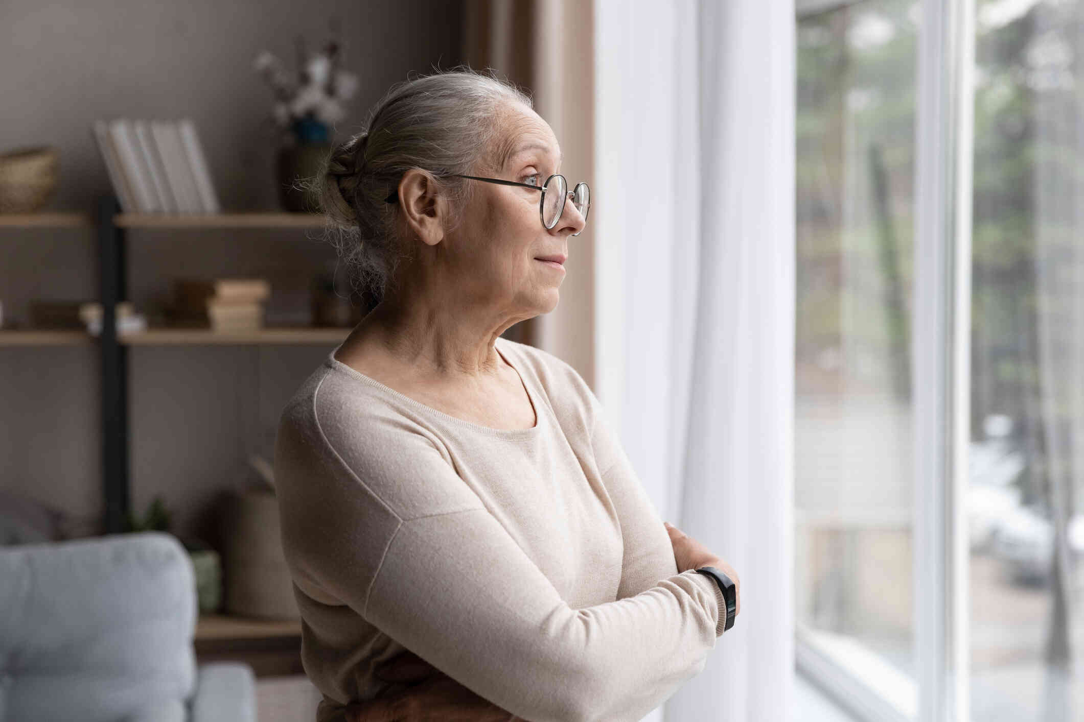 An elderly woman with glasses crosses her arms and looks out of a window.