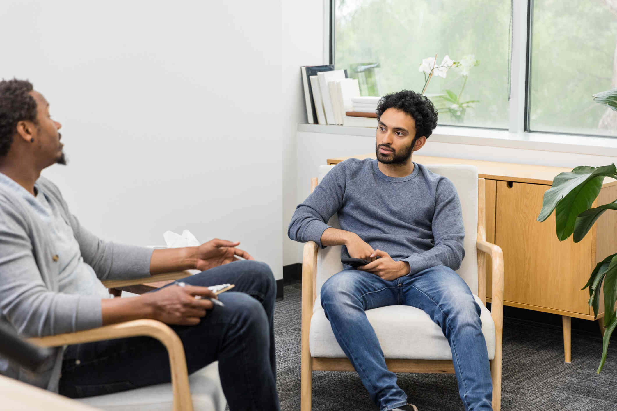 A man in a long sleeve grey shirt sits in a chair next to his male therapist as they talk during a therapy session.