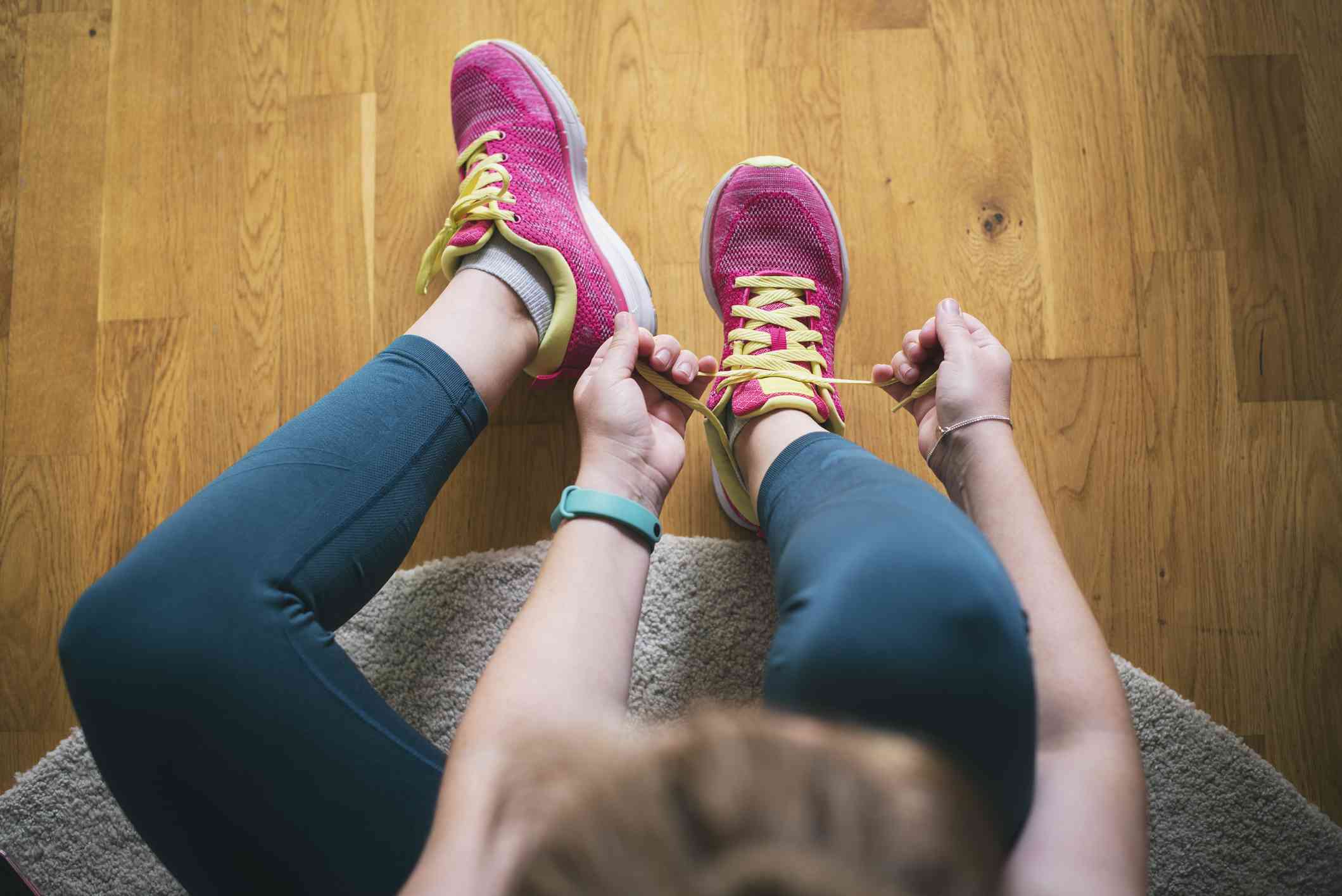 A woman wearing pink running shoes sitting on the floor while tying her shoelace