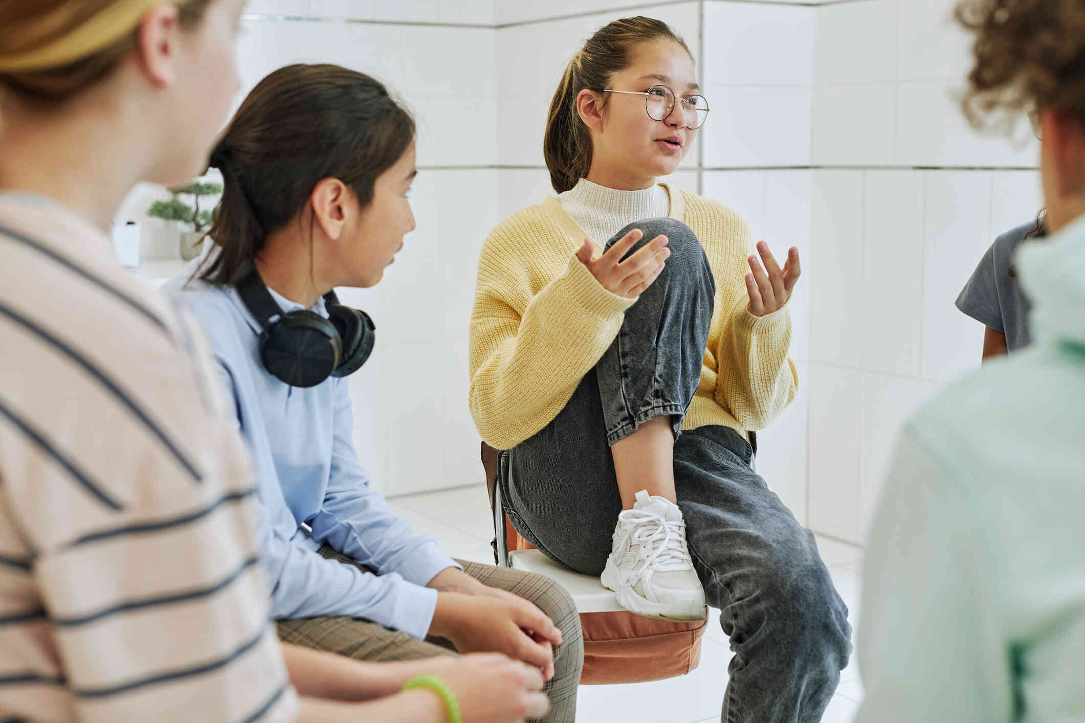 A young girl wearing glasses and a yellow sweater sits on a stool talking to a group of group of teenage girls sitting around