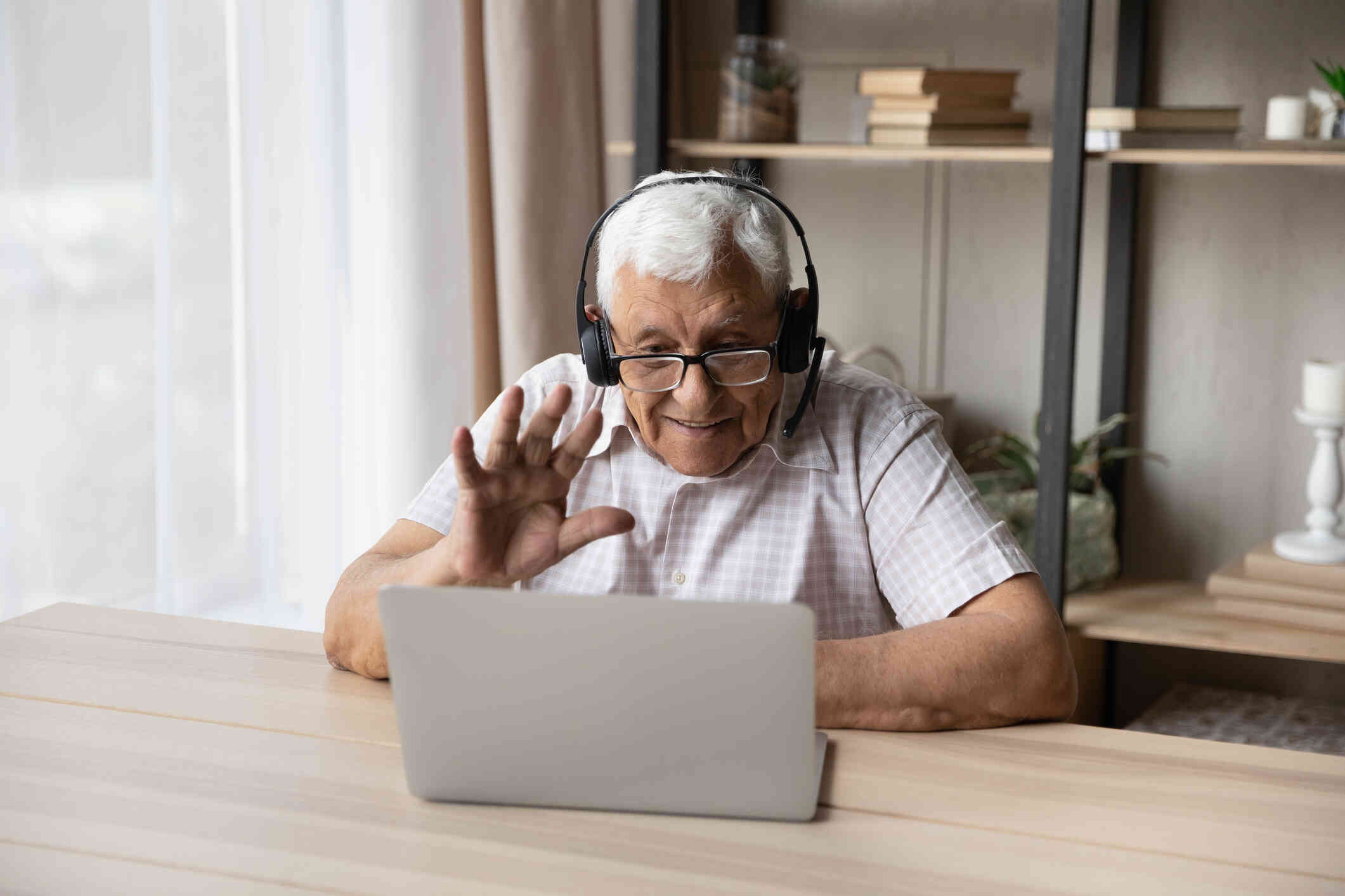 An elderly man with headphones, seated at a desk, waves and smiles at his laptop.