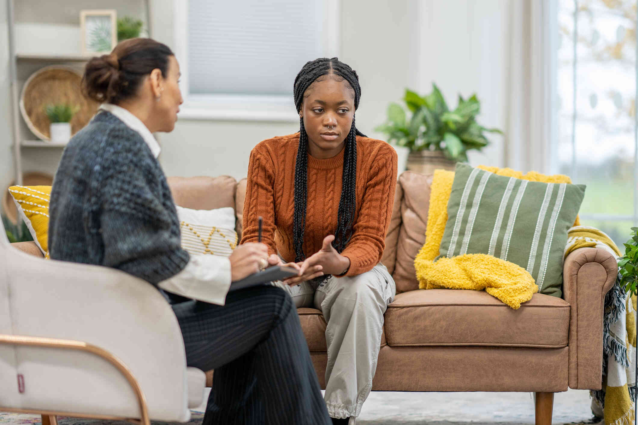 A woman in an orange sweater means over sadly while sitting on a couch and talksing to the female therapist sitting across from her.