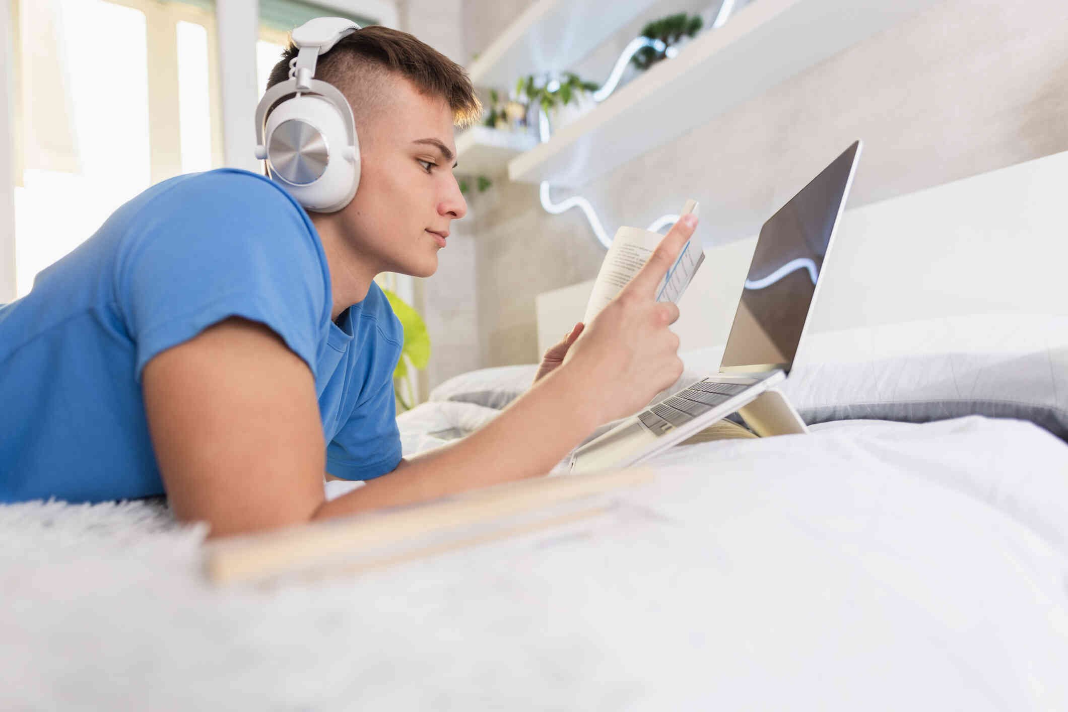 A young man wearing headphones lying on a bed and reading a book in front of his laptop.