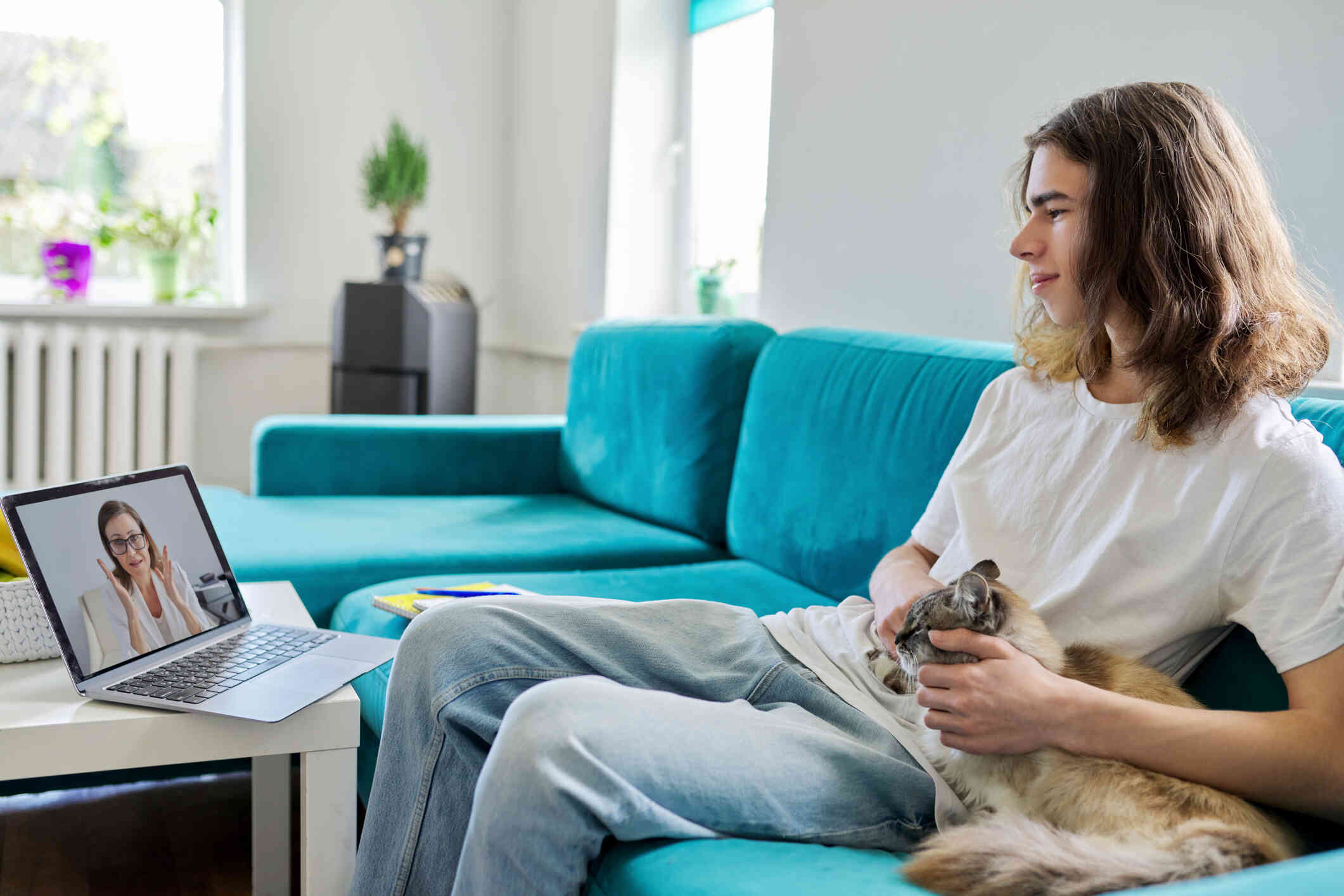 A young man with long hair and a white t-shirt sits on a blue counch with a brown cat next to him while focusing on a laptop sitting on the table in front of him. A woman speaks to him through the screen.