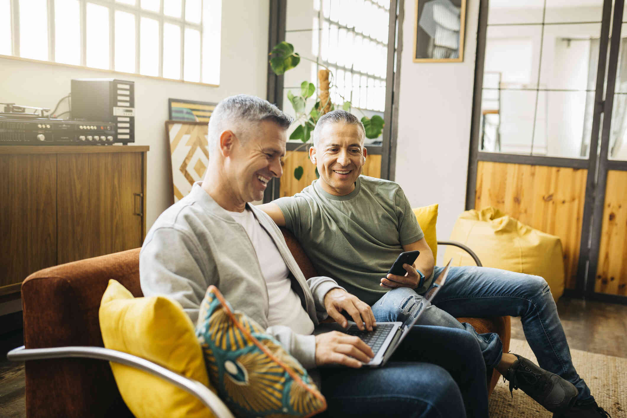 Two middle aged male friends sit next to each other on the couch with their laptop and phone while smiling.