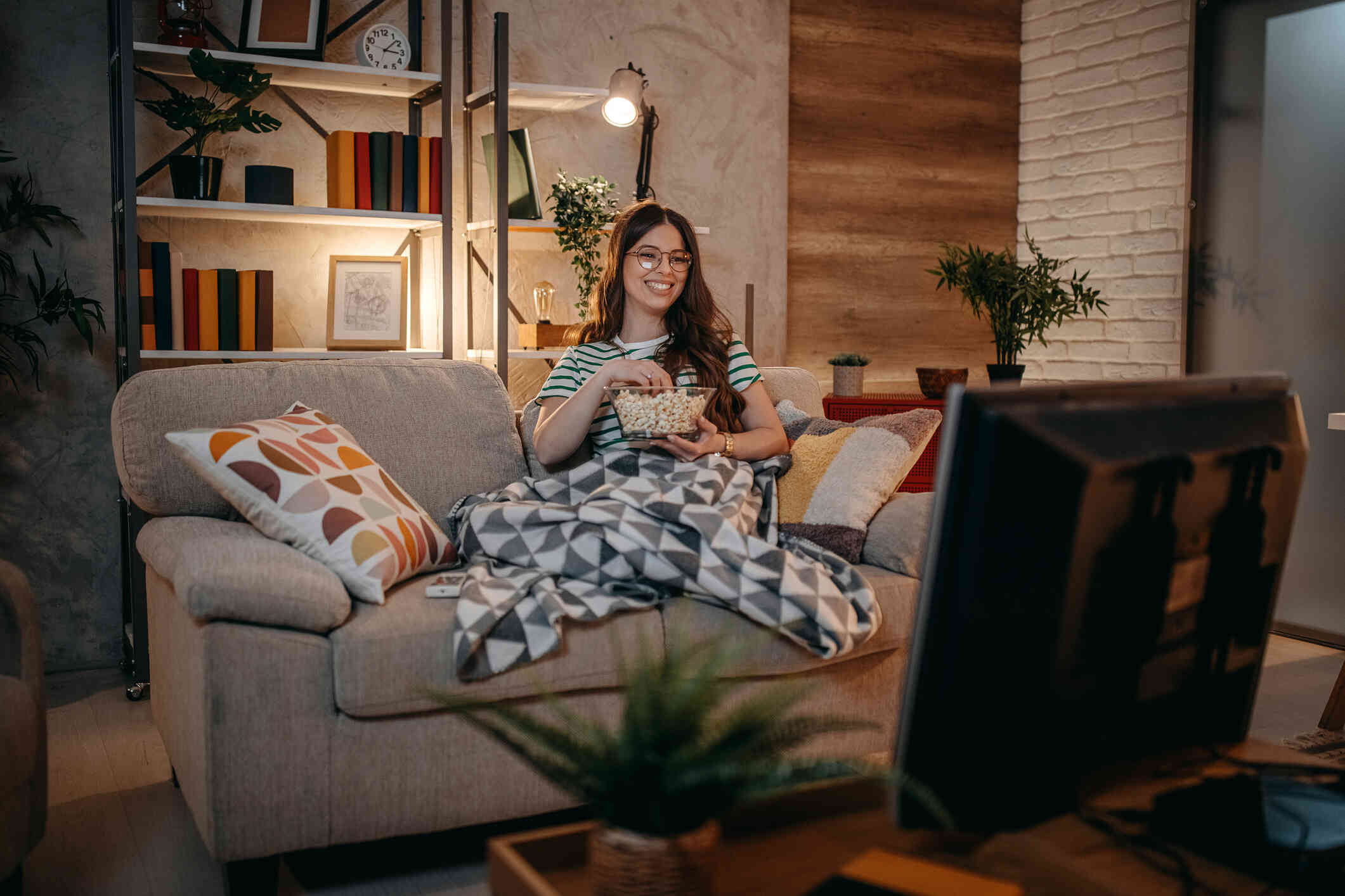 A woman smiles as she sits on a couch with a blanket and eats popcorn while watching television.