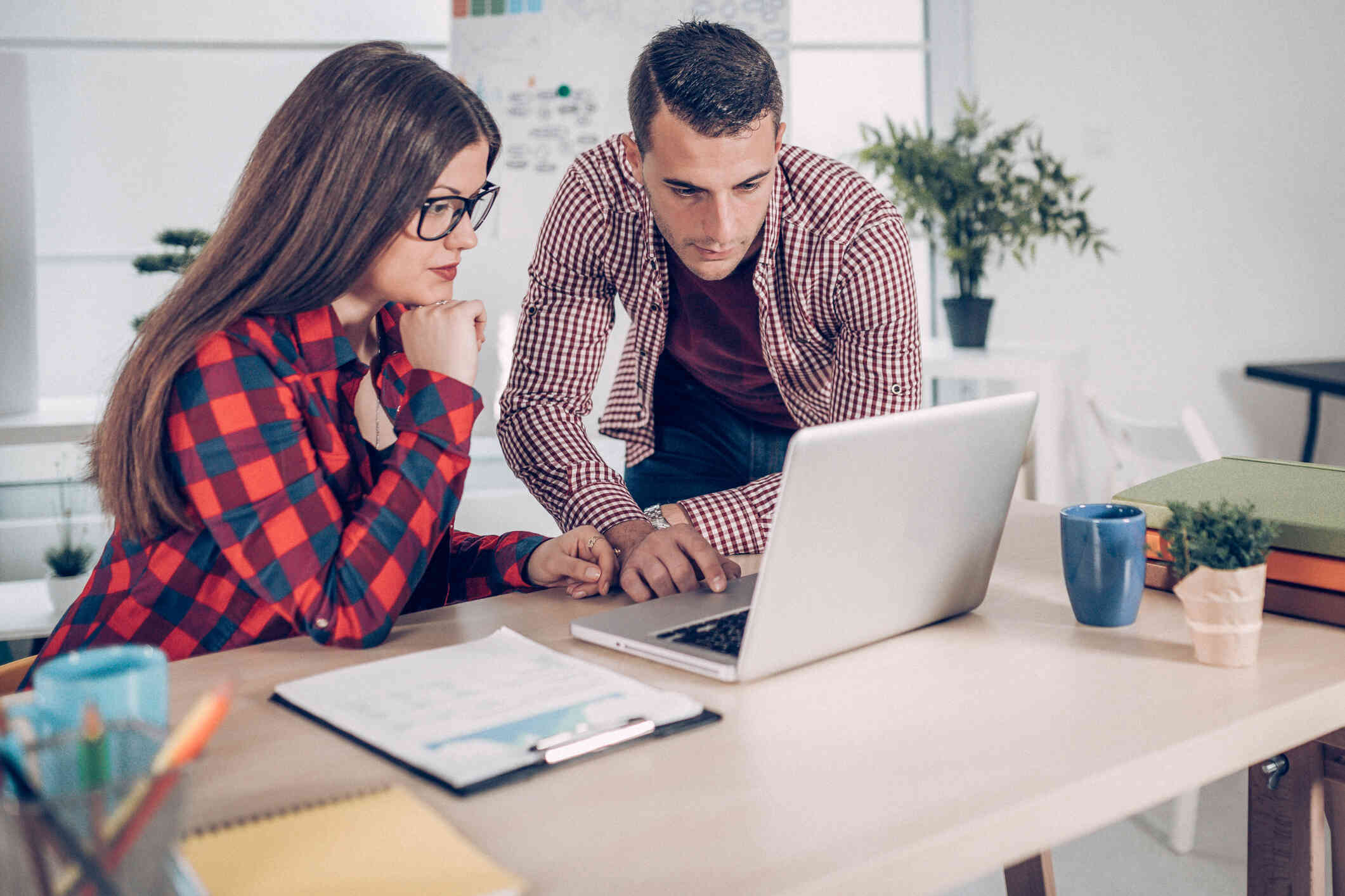 A couple sits at a desk, both busy searching for something on the laptop.