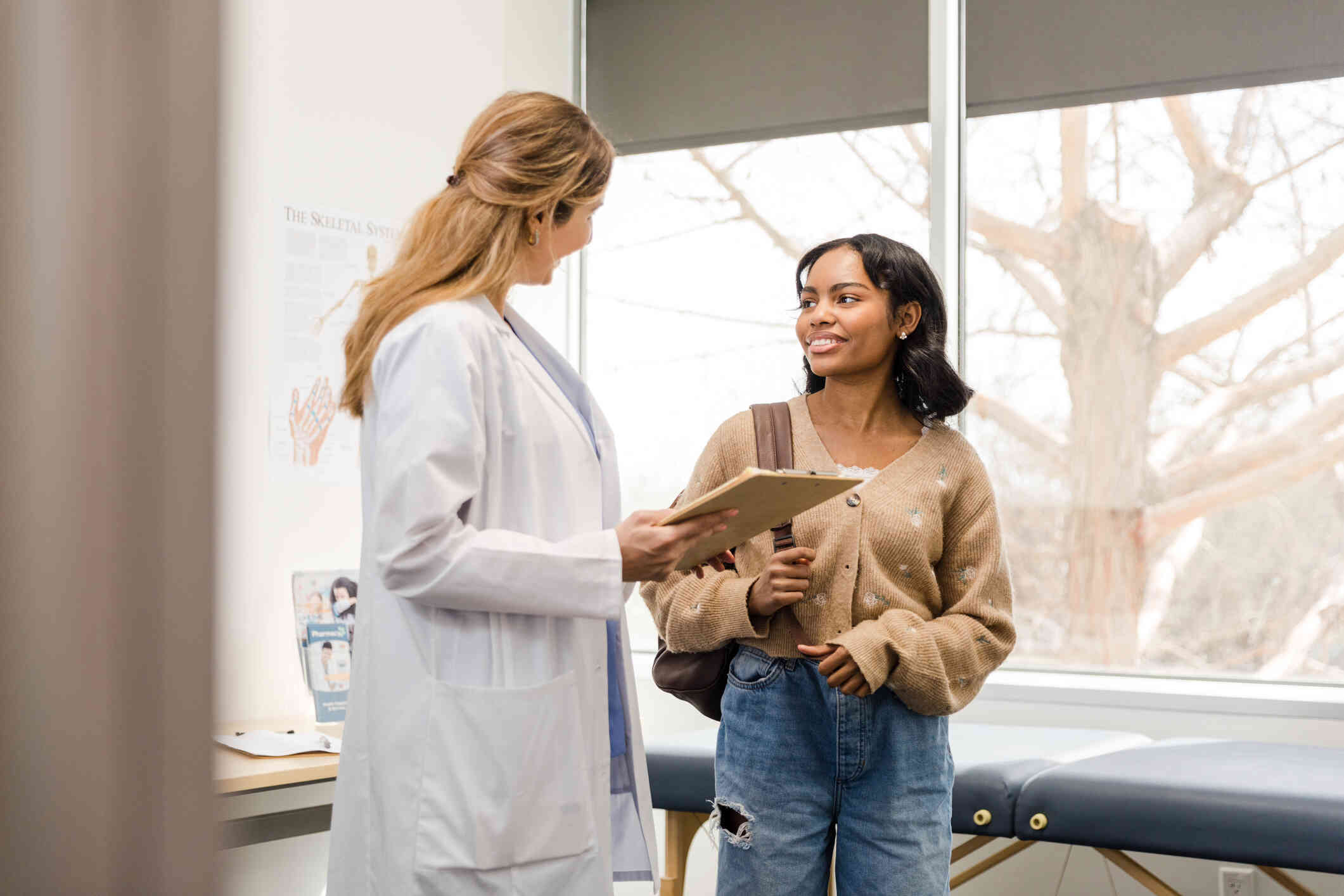 A young woman wearing a brown sweater and a doctor in a white coat holding a clipboard are in a doctors office while having a conversation.