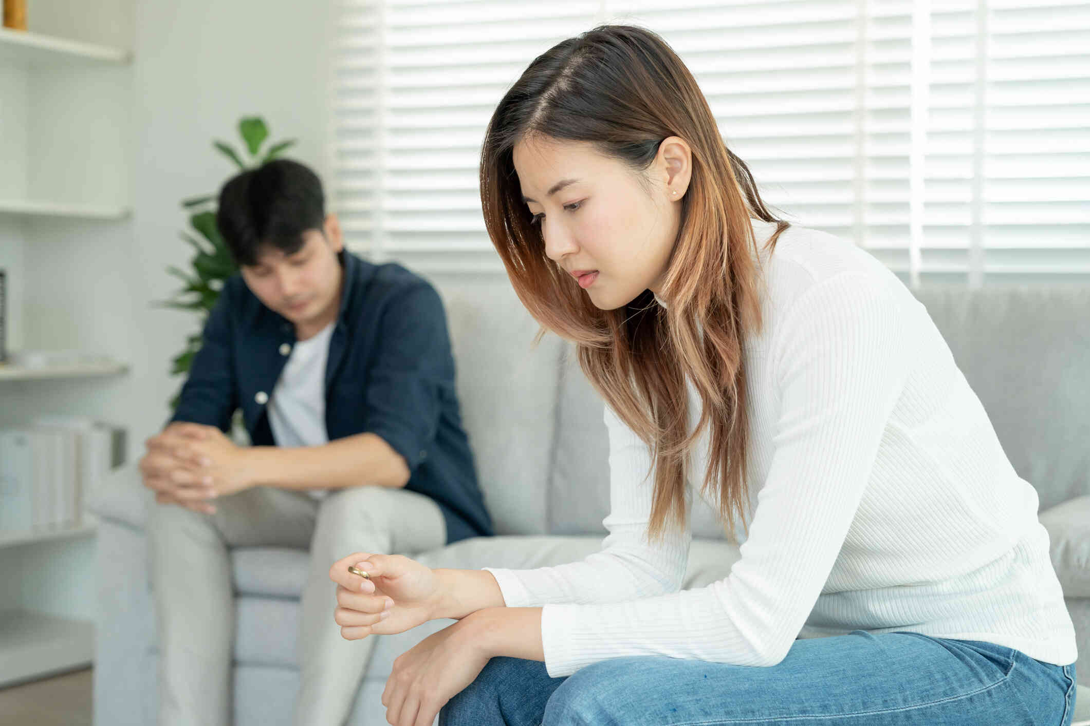 A woman in a white shirt sits sadly on the couch while gazing down as her male parter sits in the background with a sad expression.