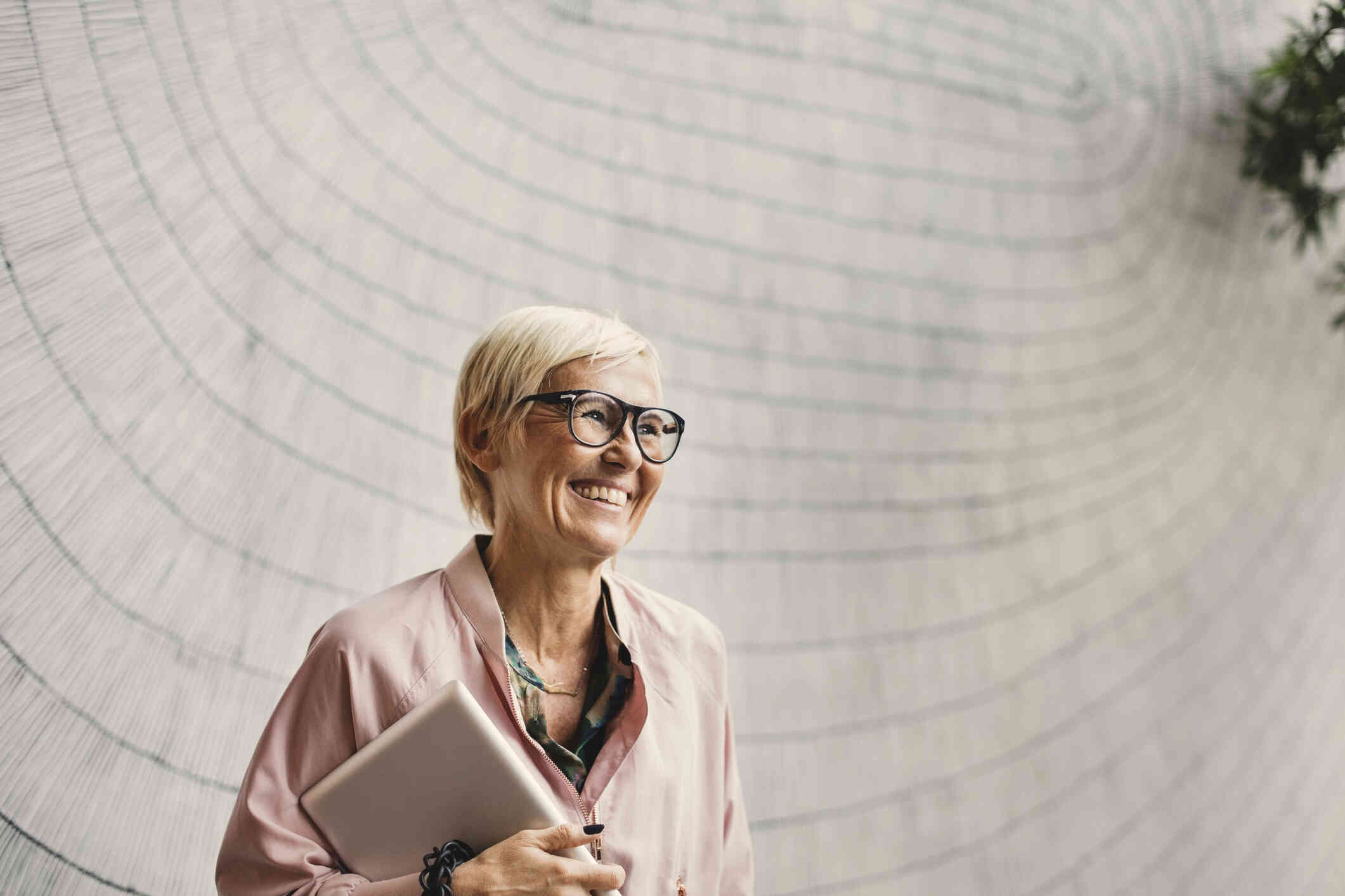 A woman with short hair and glasses smiles as she holds a laptop under her arm and stands in a marble building.