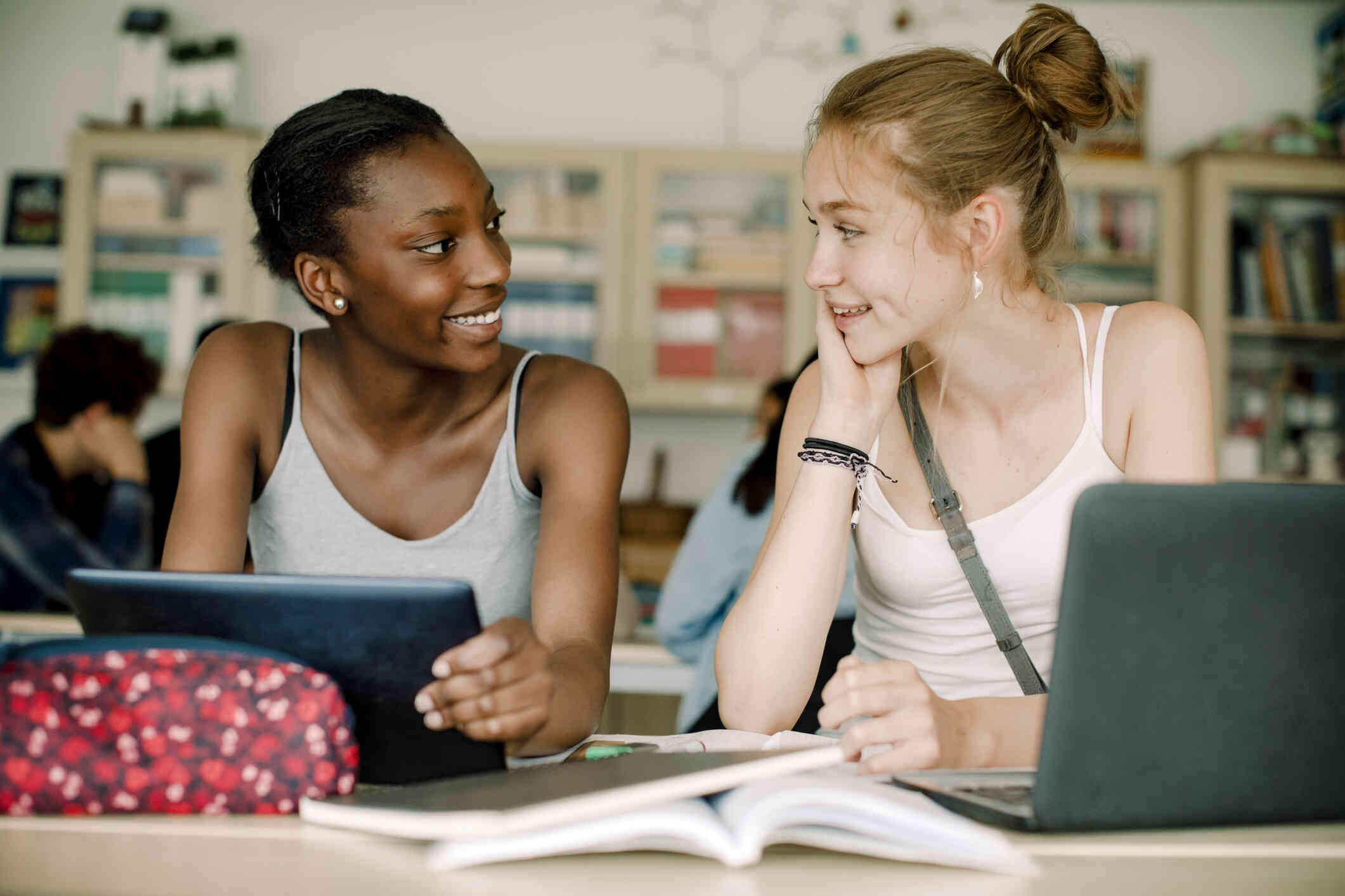Two teen girls sit side by side at a table with notebooks and thair laptops while they smile at one another.