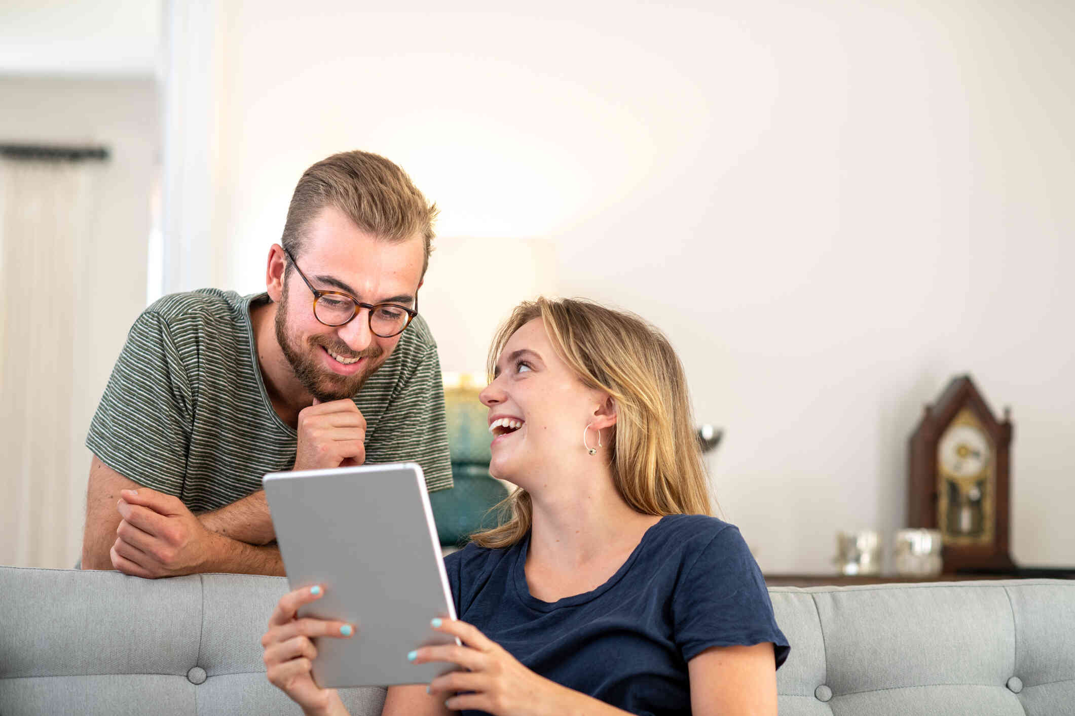 A woman sits on a couch and smiles as she holds a tablet. A man with glasses leans down next to her smiling as he looks at the tablet.