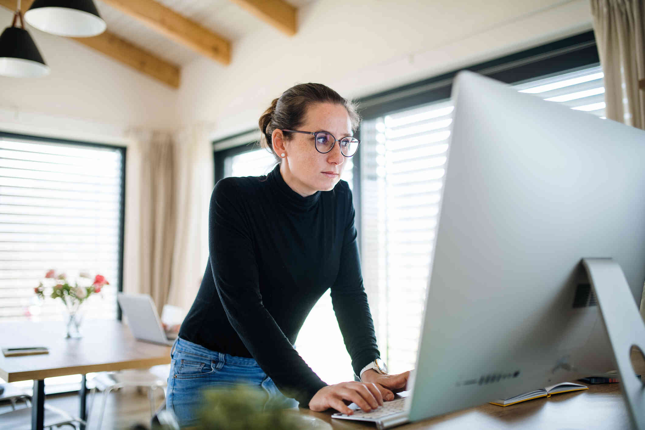 A woman in a black shirt and glasses sits at a counter in her home infront of her computer while typing with a serious expression.