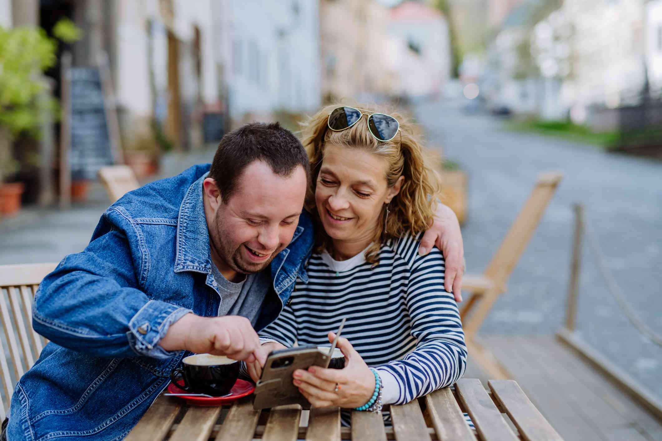 A man and a woman happily look at a smartphone together while sitting at an outdoor café.