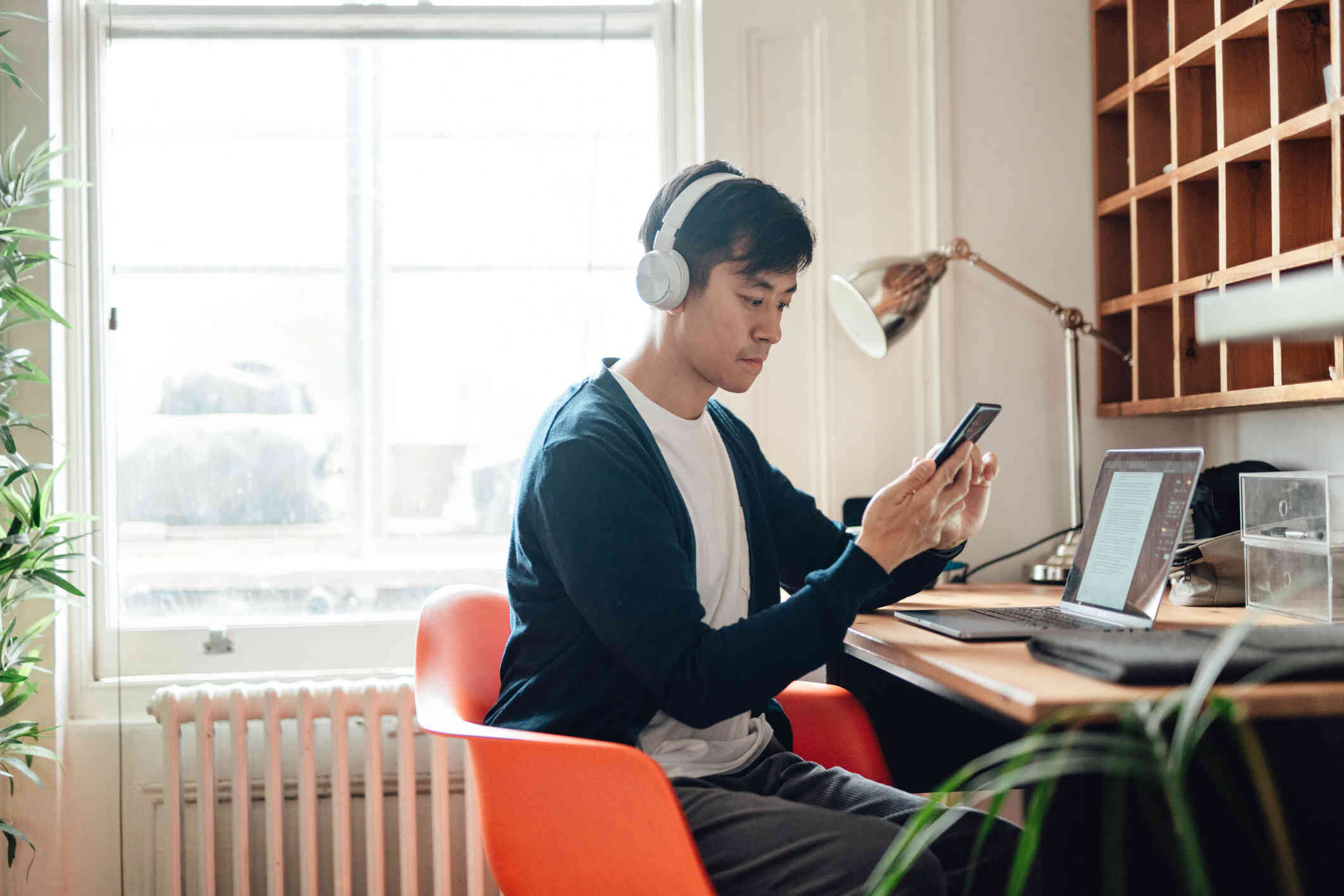 A man in ablue sweater sits at his computer desk while wearing white headphone and looking at the phone in his hands with a serious expression.