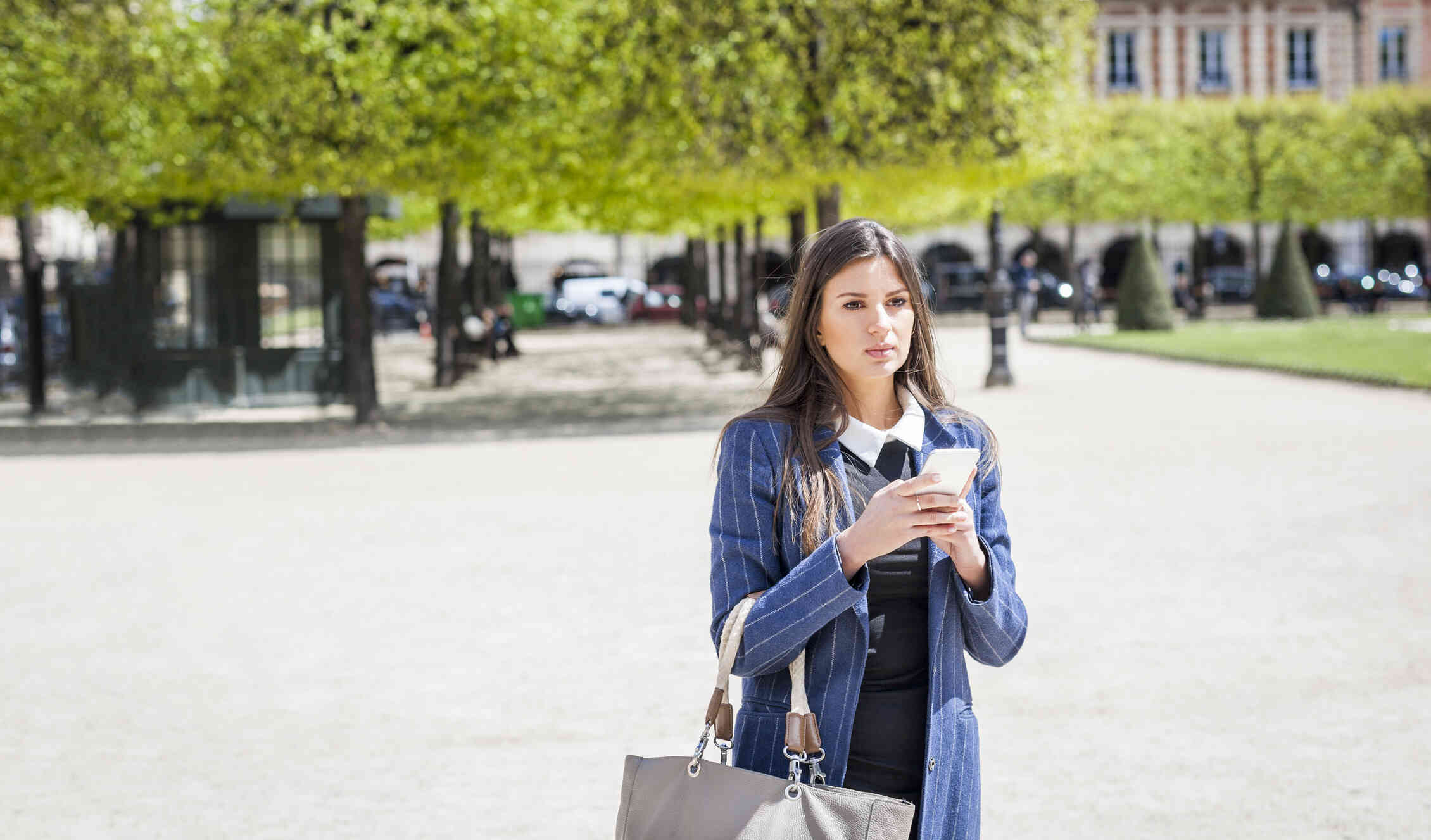A woman in a blue coat walks outside on a very sunny day while carrying her large purse and holding her cellphone in her hands.
