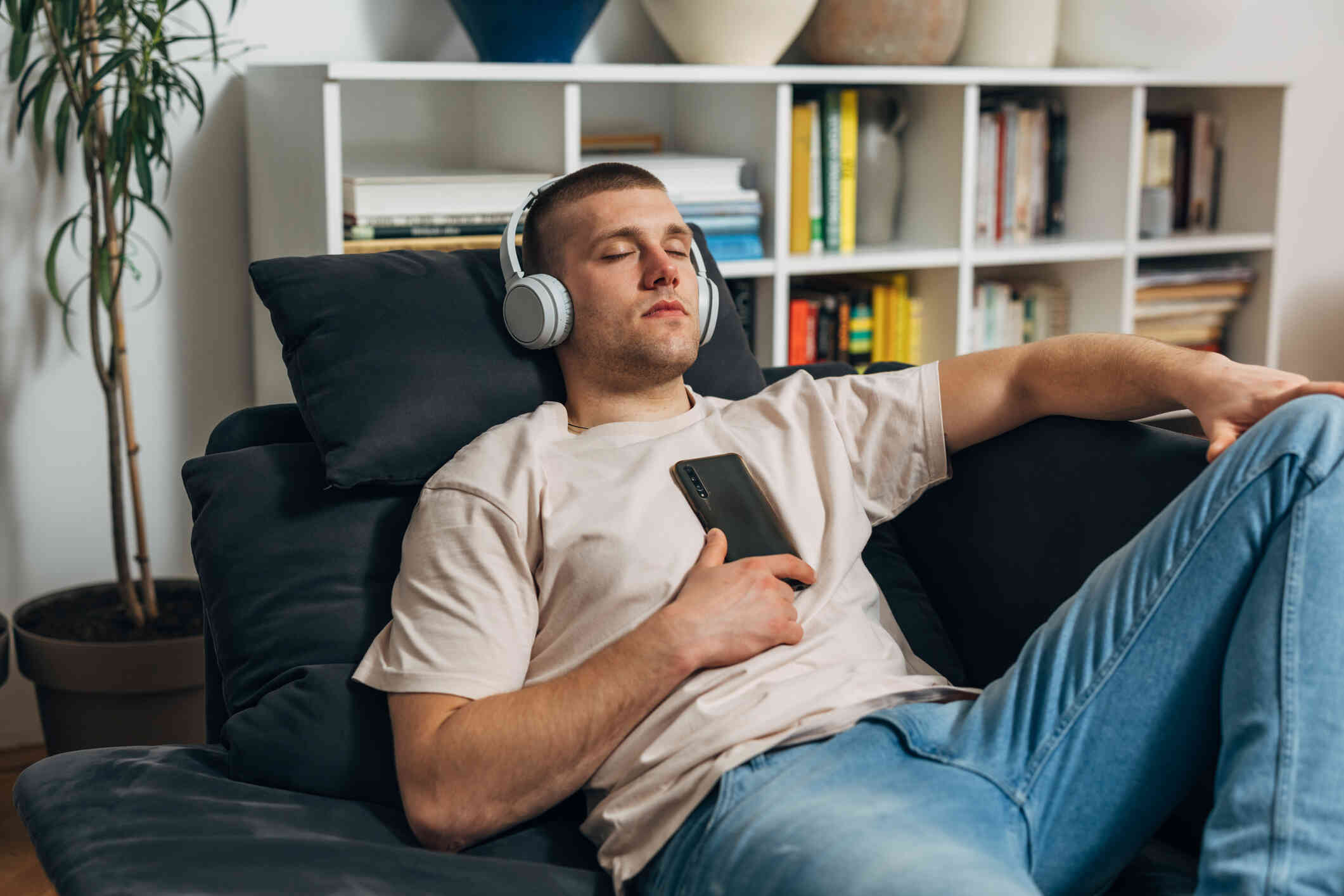 A man reclines on his black couch with his eyes closed while wearing white headphones and resting a phone on his chest.