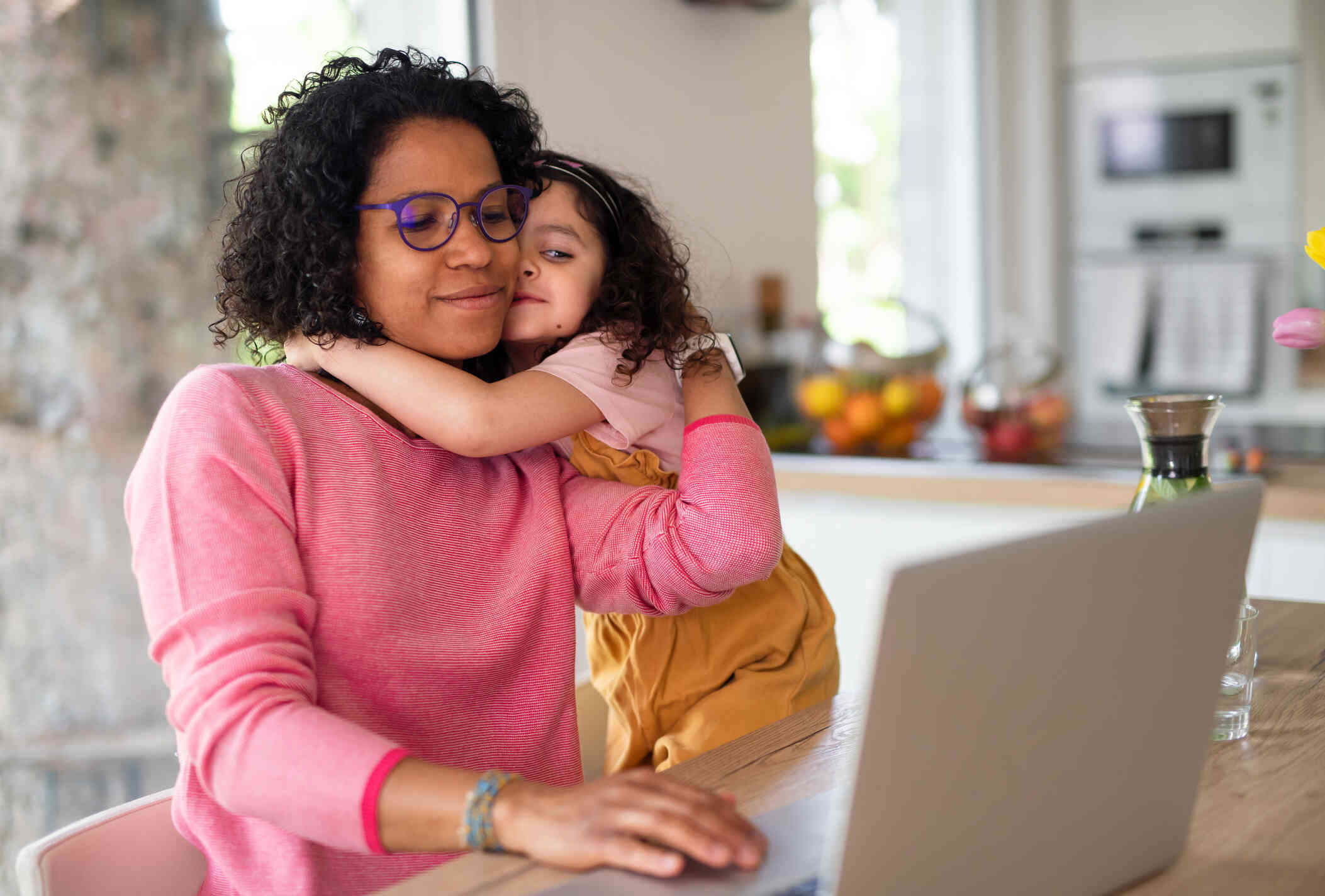 A young girl hugs her mother around the neck as the mother sits at the kitchen table with her laptop open on the table infront of her.