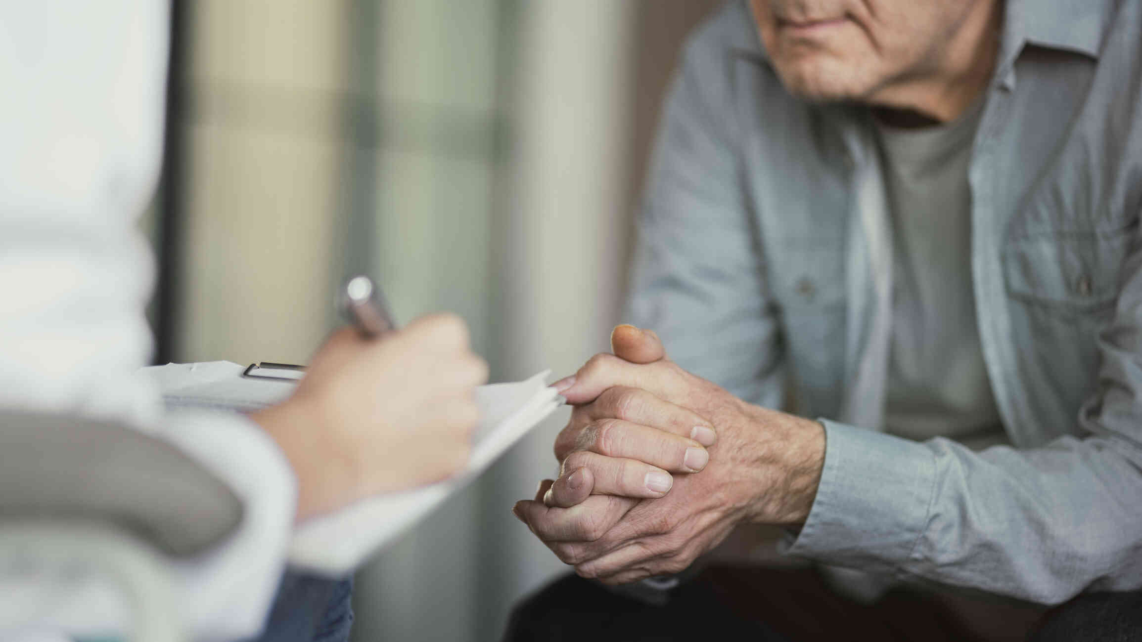 A close up of a man leaning forward in his chair and clasping his hands together as the therapist sitting across frm him takes notes on a clipboard.