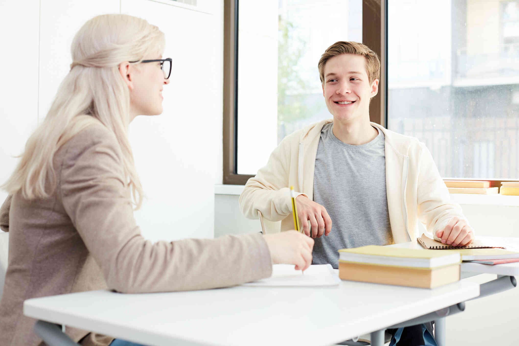 A young man smiles as he sits across from a woman with glasses at a desk with notebooks out in front of them.