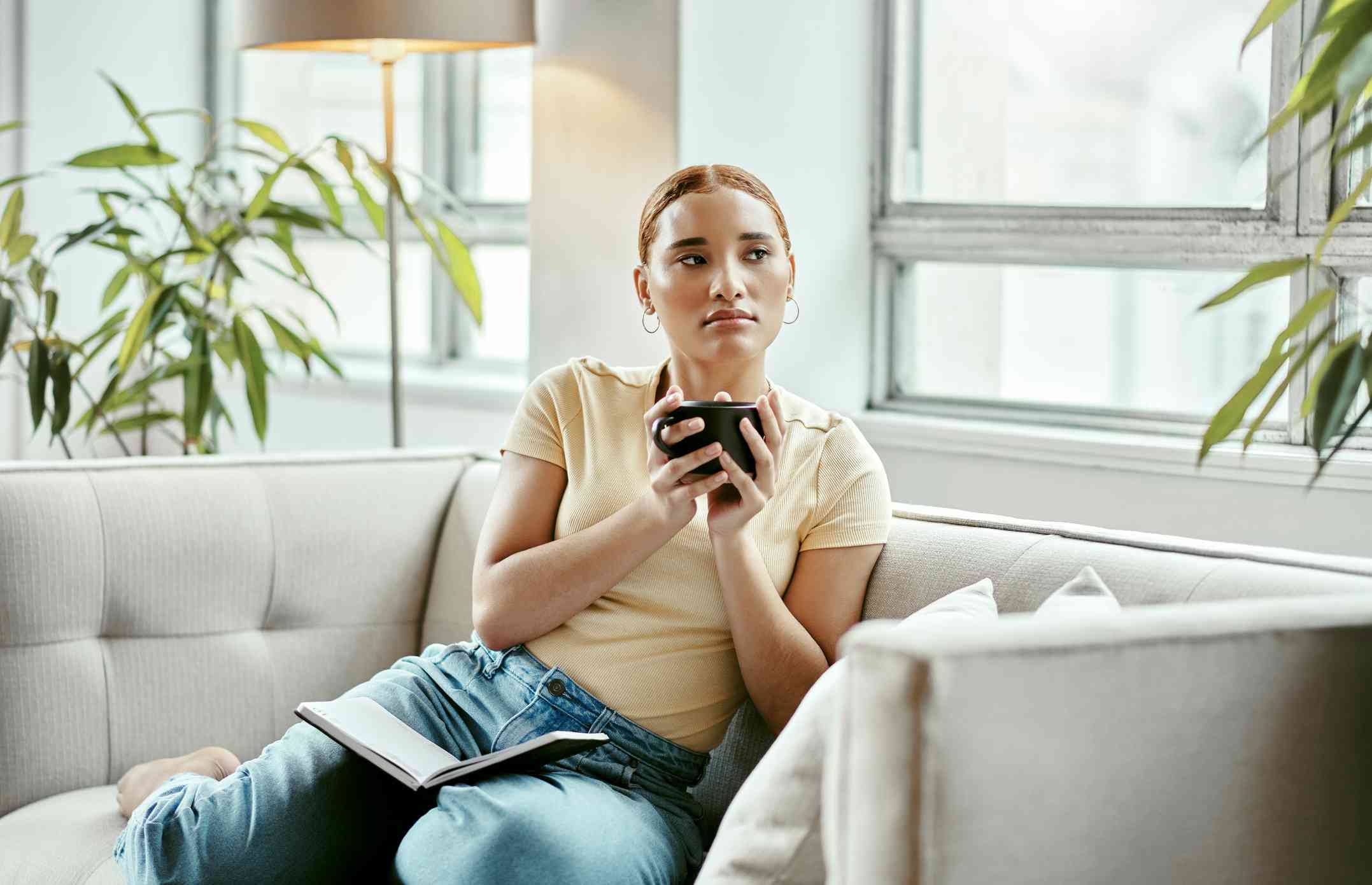 A woman in a yellow shirt sits on the couch with a coffe mug in her hands and a book open in her lap as she gazes off sadly.