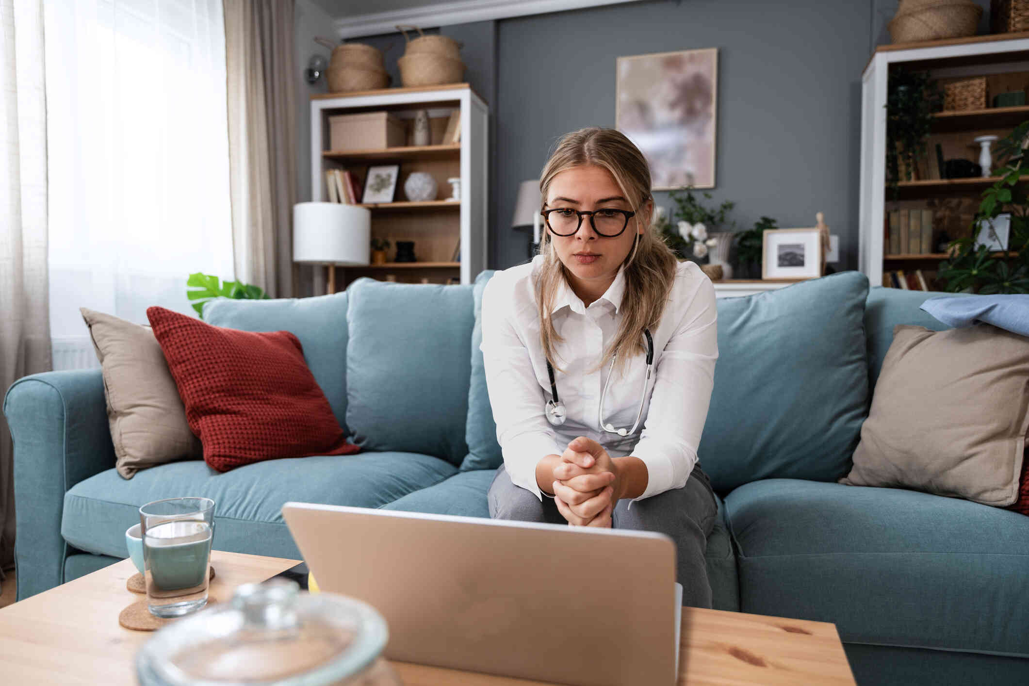 A young female in glasses, seated on a blue couch, looks attentively at her laptop.