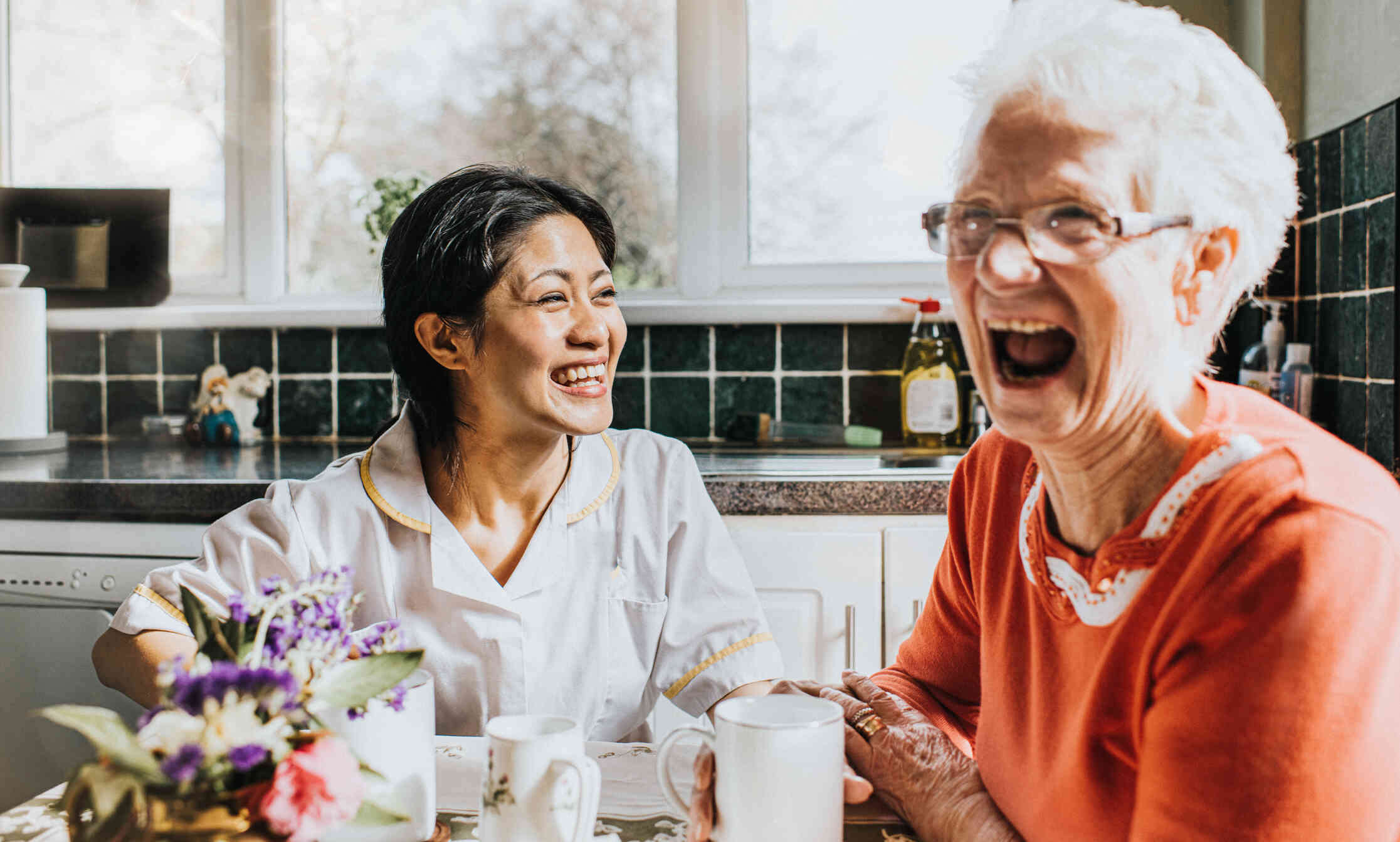 A woman in awhite shirt sits at the kitchen table with her elderly female relative as they laugh together on a sunny morning.