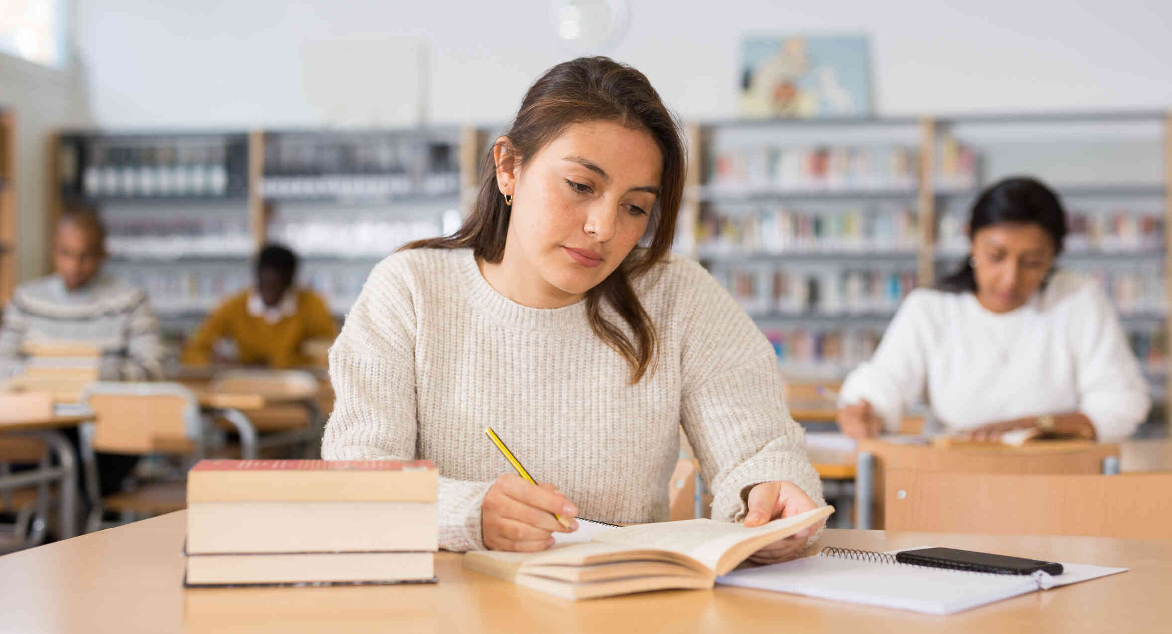 A woman in a tan sweater sits at a table in the library and writes in a notebook while looking at a book.