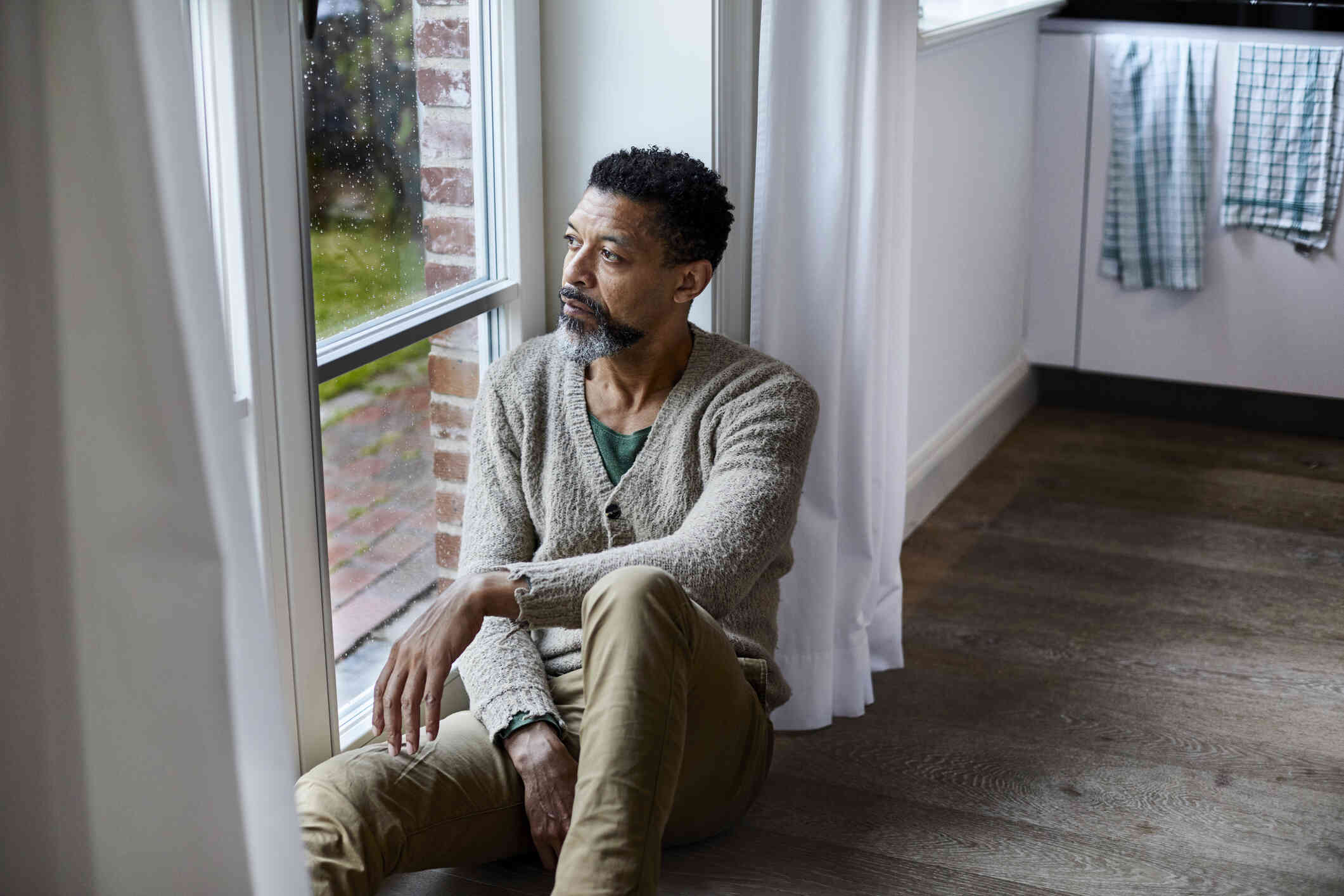 A middle aged man in a grey sweater sits on the floor of his home near a wondow and gazes out while deep in thought.