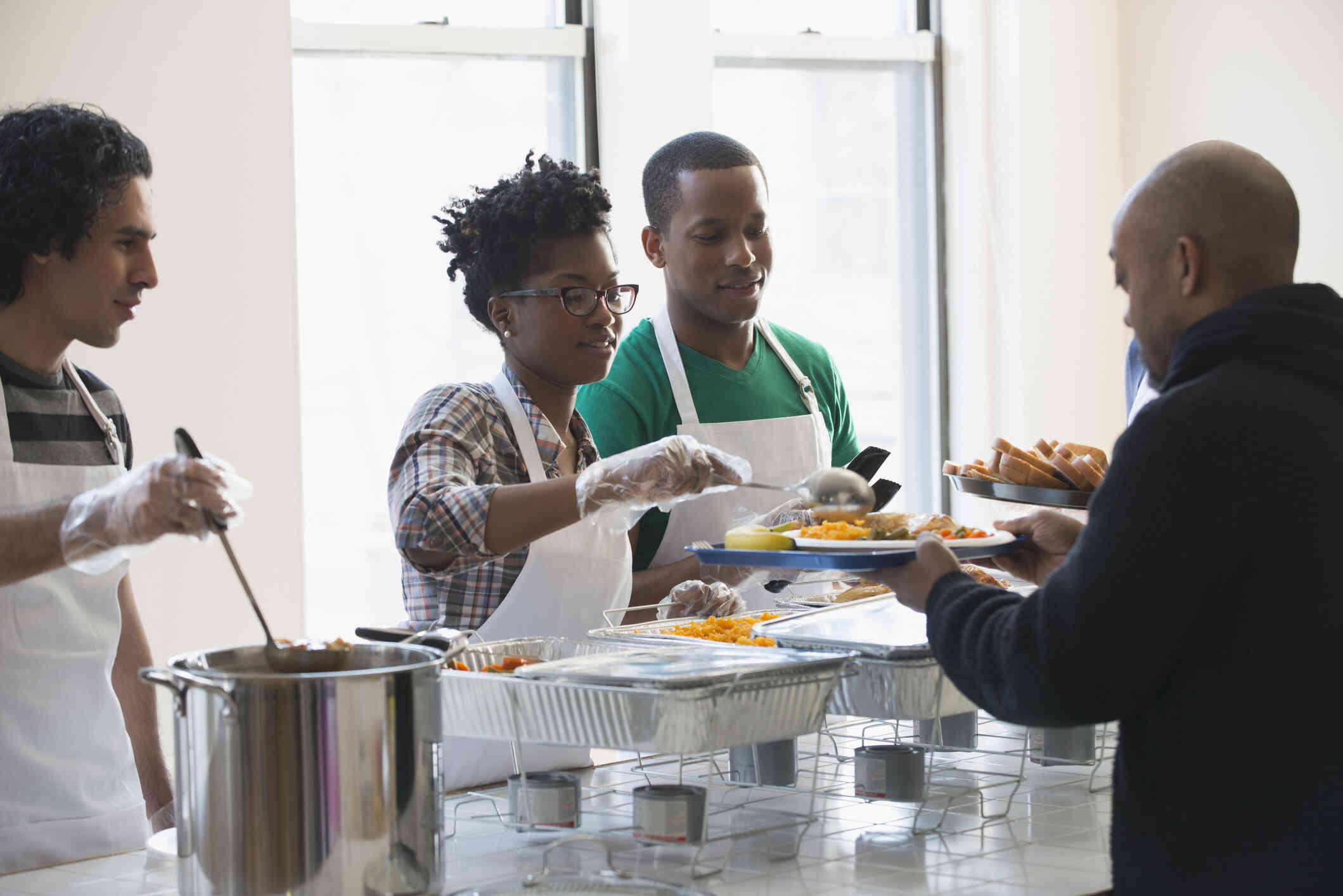 A woman wearing an apron and gloves serves food to a man who is waiting in line. Two men stand beside the woman ready to serve food our of trays and pots to the next people in line.