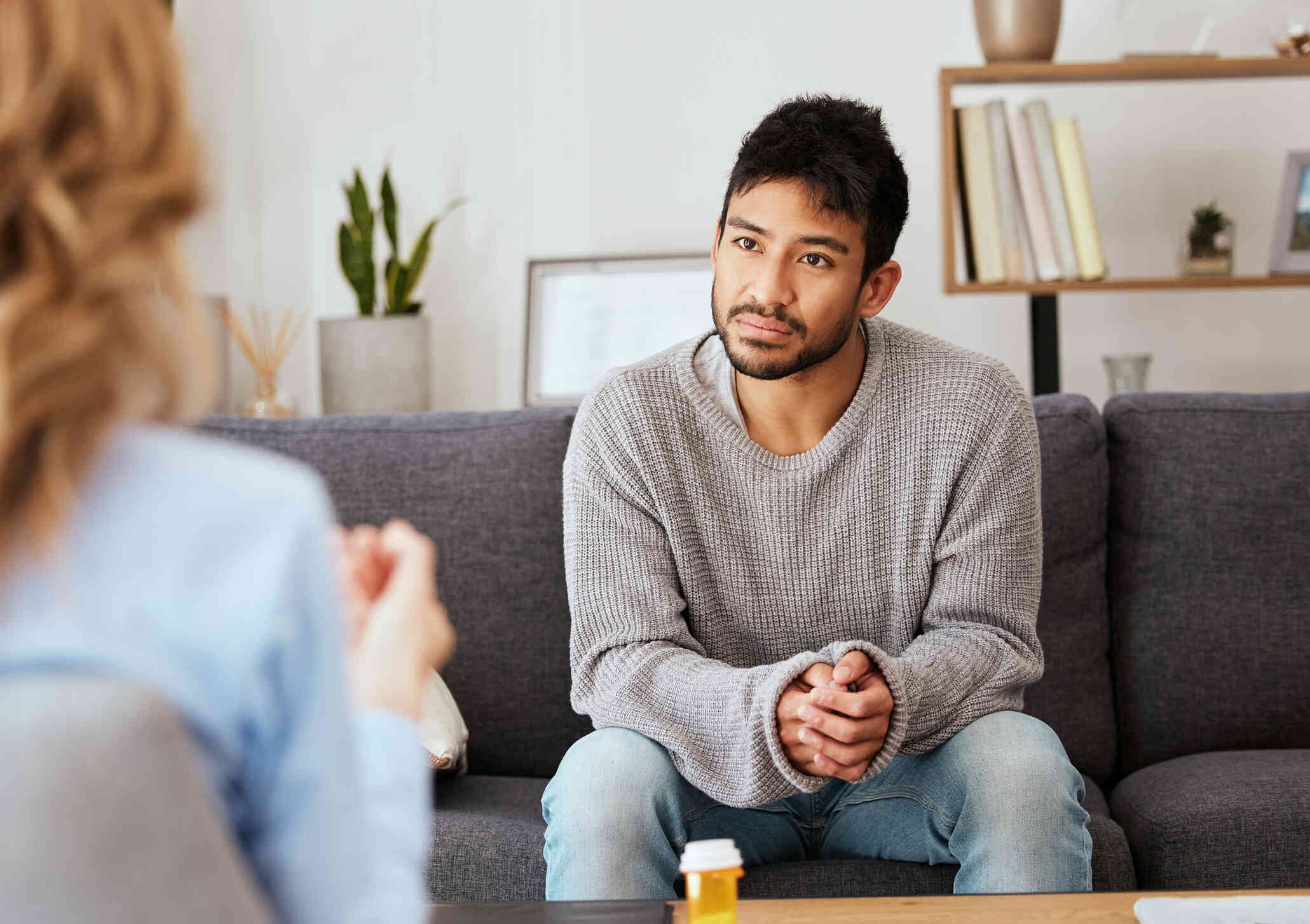 A man in a grey sweater sits on a couch and leans forward with his hands clasped together and aworried expression on his face as he listens to the female therapist sitting across from him.