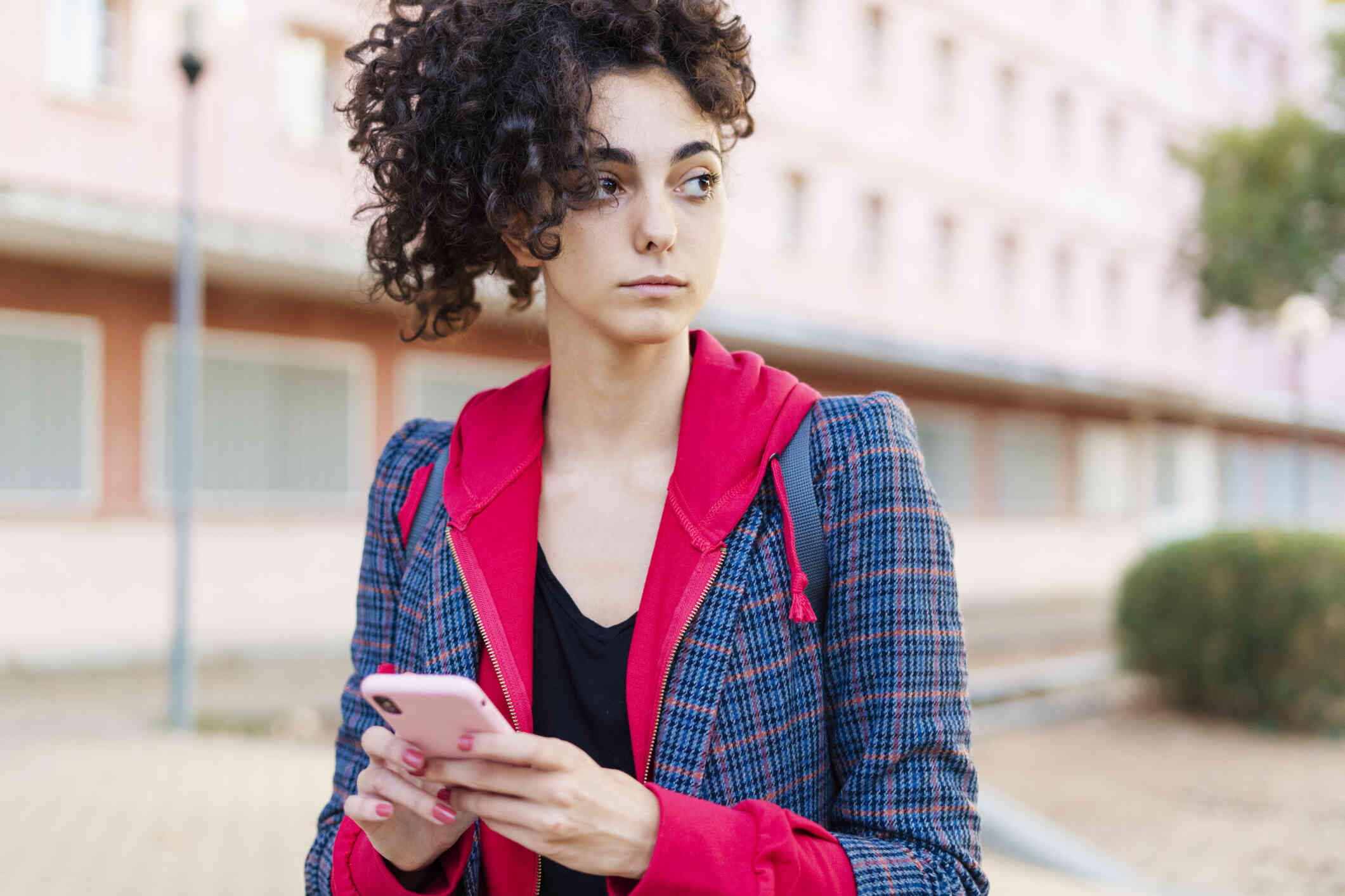 A woman in a blue blazer and red sweater stands outside while holding her phone in her hand and gazes off with a blank expression.