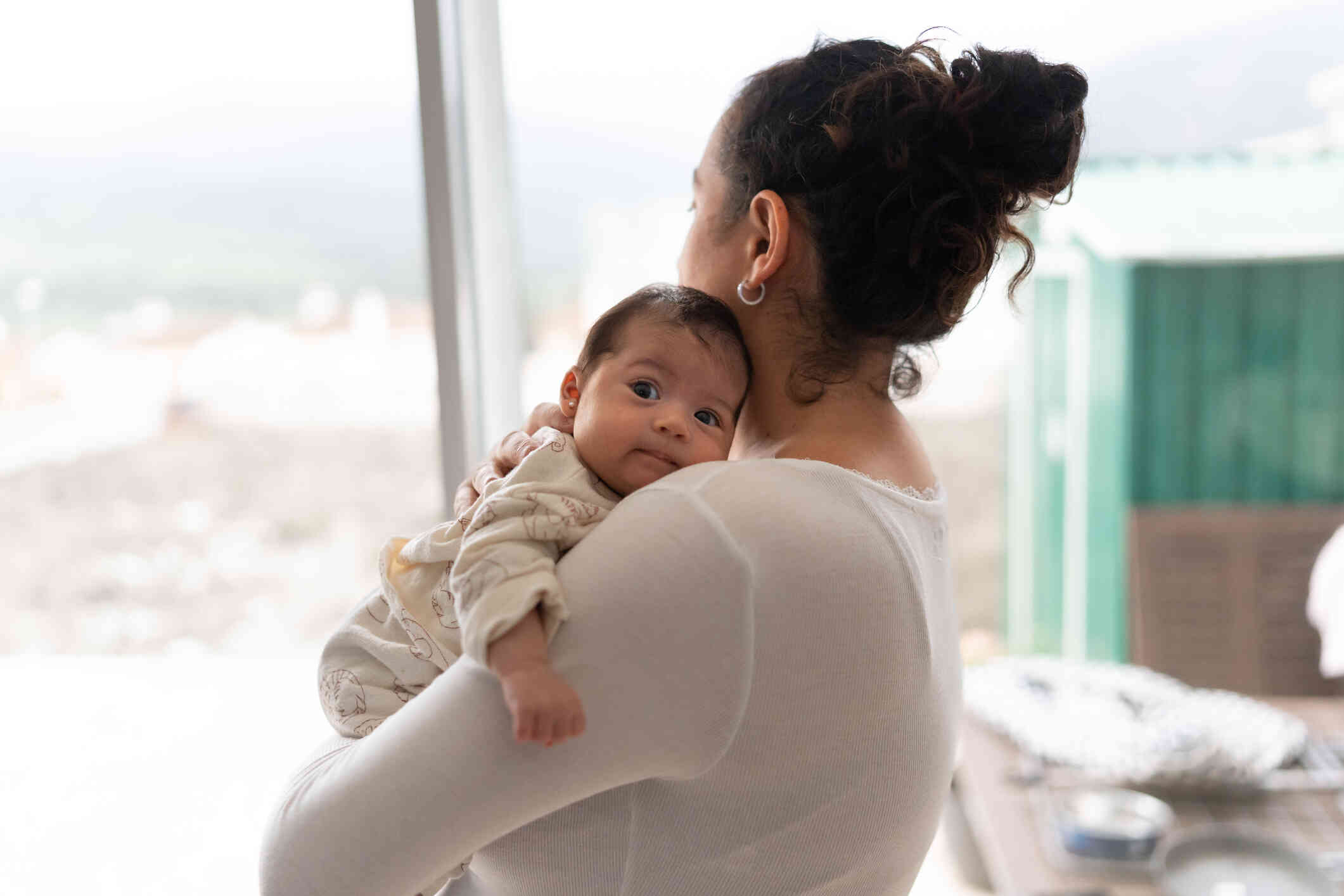 A mother in a white shirt stand in her home and holds her newborn infant on her chest.