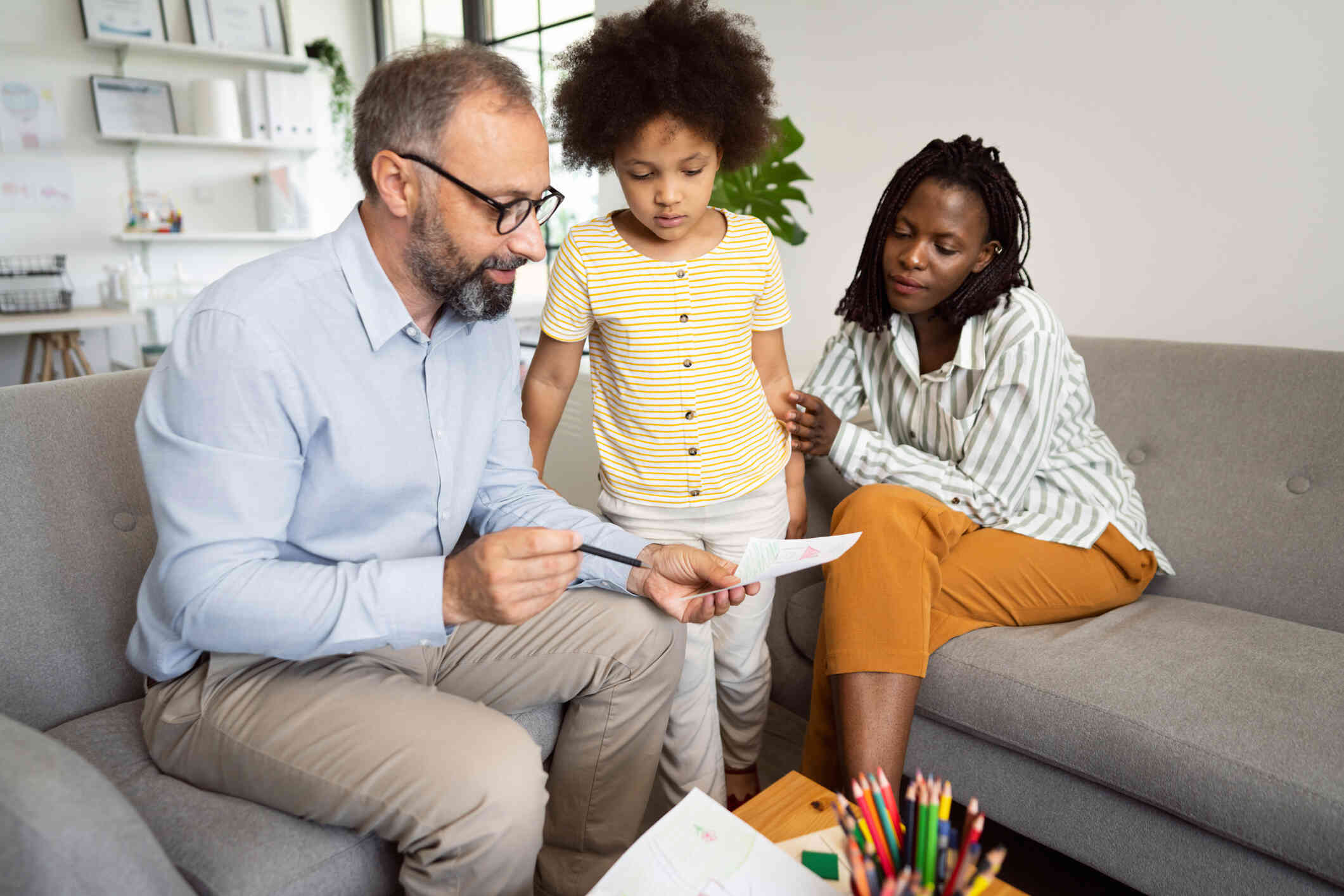 A young girl in a yellow striped shirt stands next to her parents sitting on the couch while looking at drawings on paper