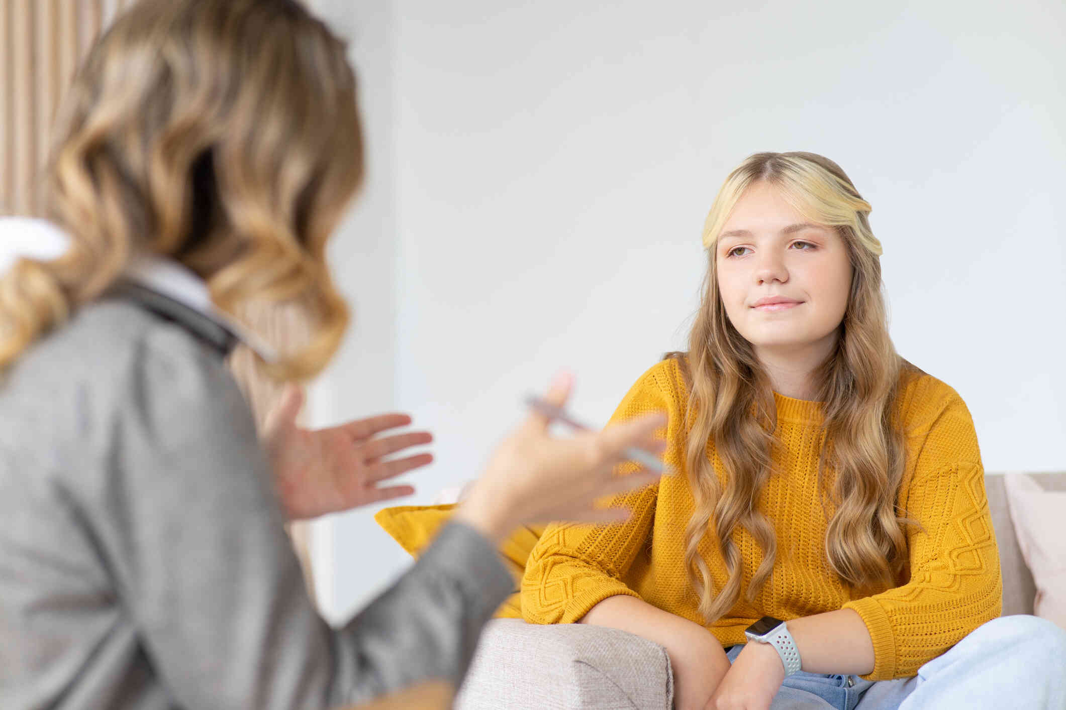 A teen girl with long blonde hair and a yellow shirt smiles slightly while looking at a woman who is sitting across from her speaking.